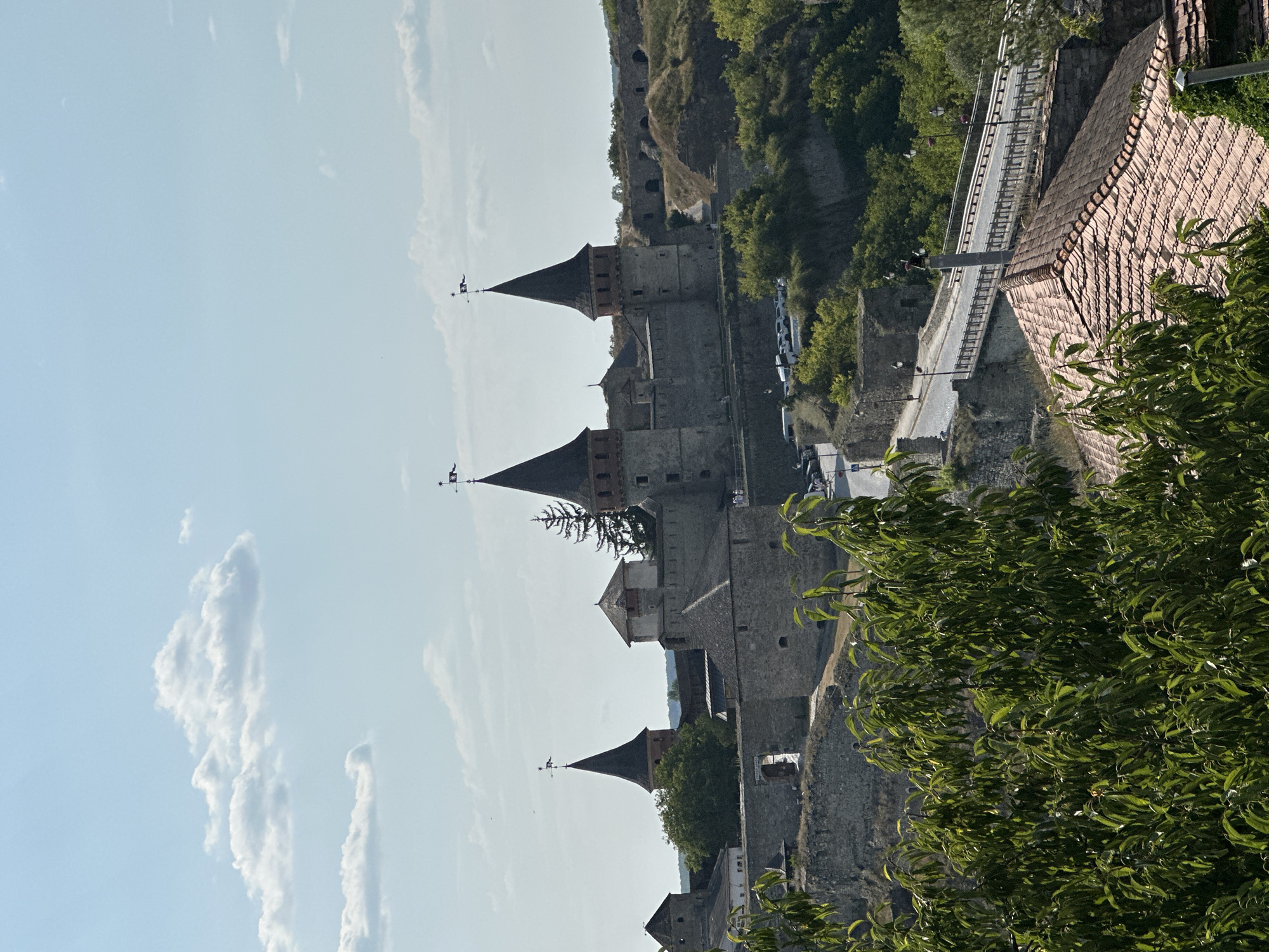 The image shows a panoramic view of the Kamianets-Podilskyi Castle in Kamianets-Podilskyi, Ukraine, on a clear, sunny day. The imposing stone castle, a historical fortress, dominates the center of the frame with its distinctive conical towers. A bridge, with cars and streetlights visible, crosses a ravine in the foreground, leading towards the castle. Lush green trees and foliage surround the castle and the surrounding landscape, suggesting a warm, possibly summer season. The sky is a pale blue with scattered white clouds. The overall scene depicts a historic landmark in a natural, somewhat rugged environment.