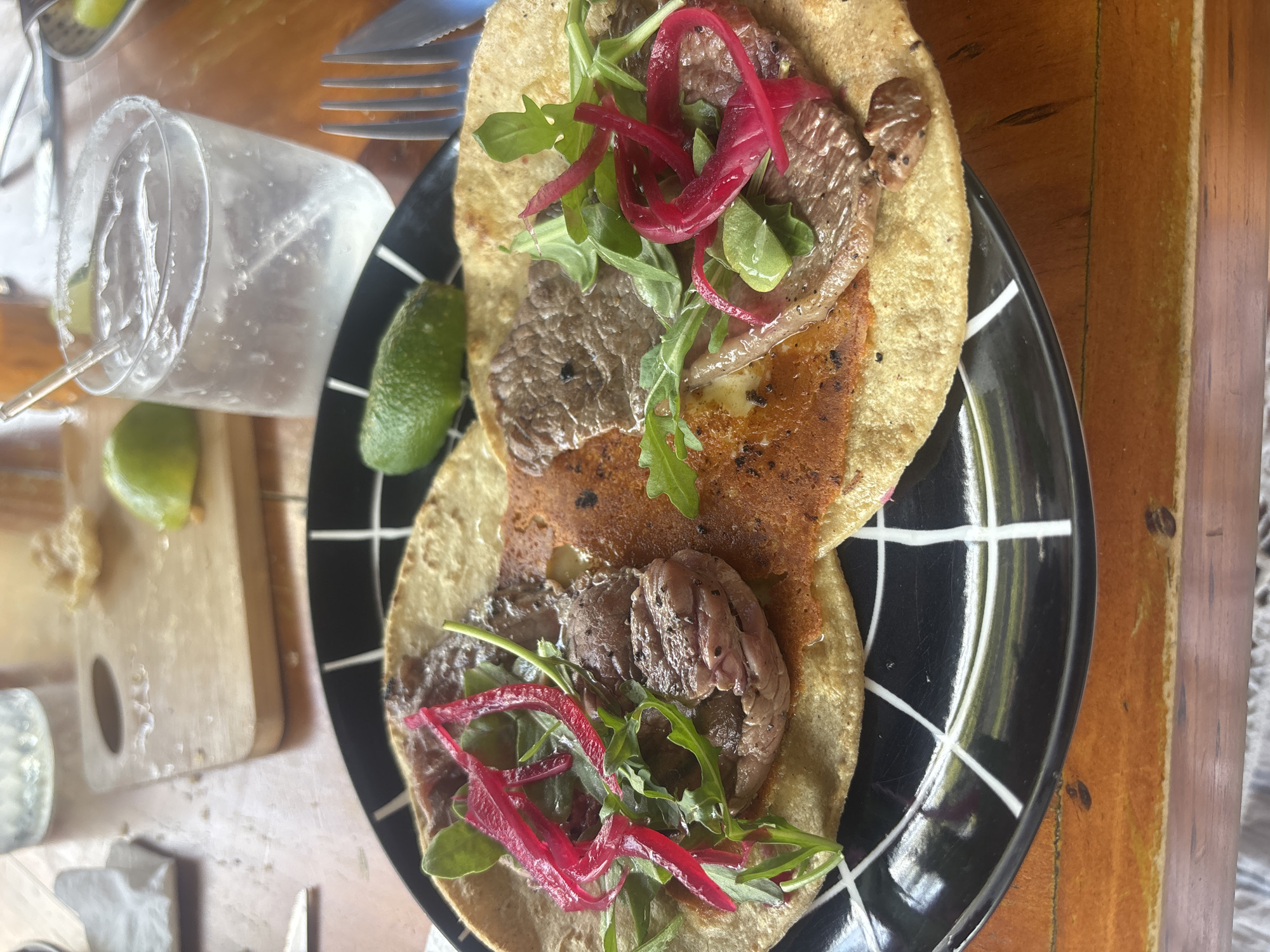The image depicts a plate of two tacos served on a black and white checkered plate, likely at a restaurant in San Andrés Cholula, Mexico. Each taco consists of a tortilla filled with sliced meat, topped with green arugula and pickled red onions. A lime wedge is placed next to the plate. A glass of iced beverage and silverware are visible on the table. The table is wooden.