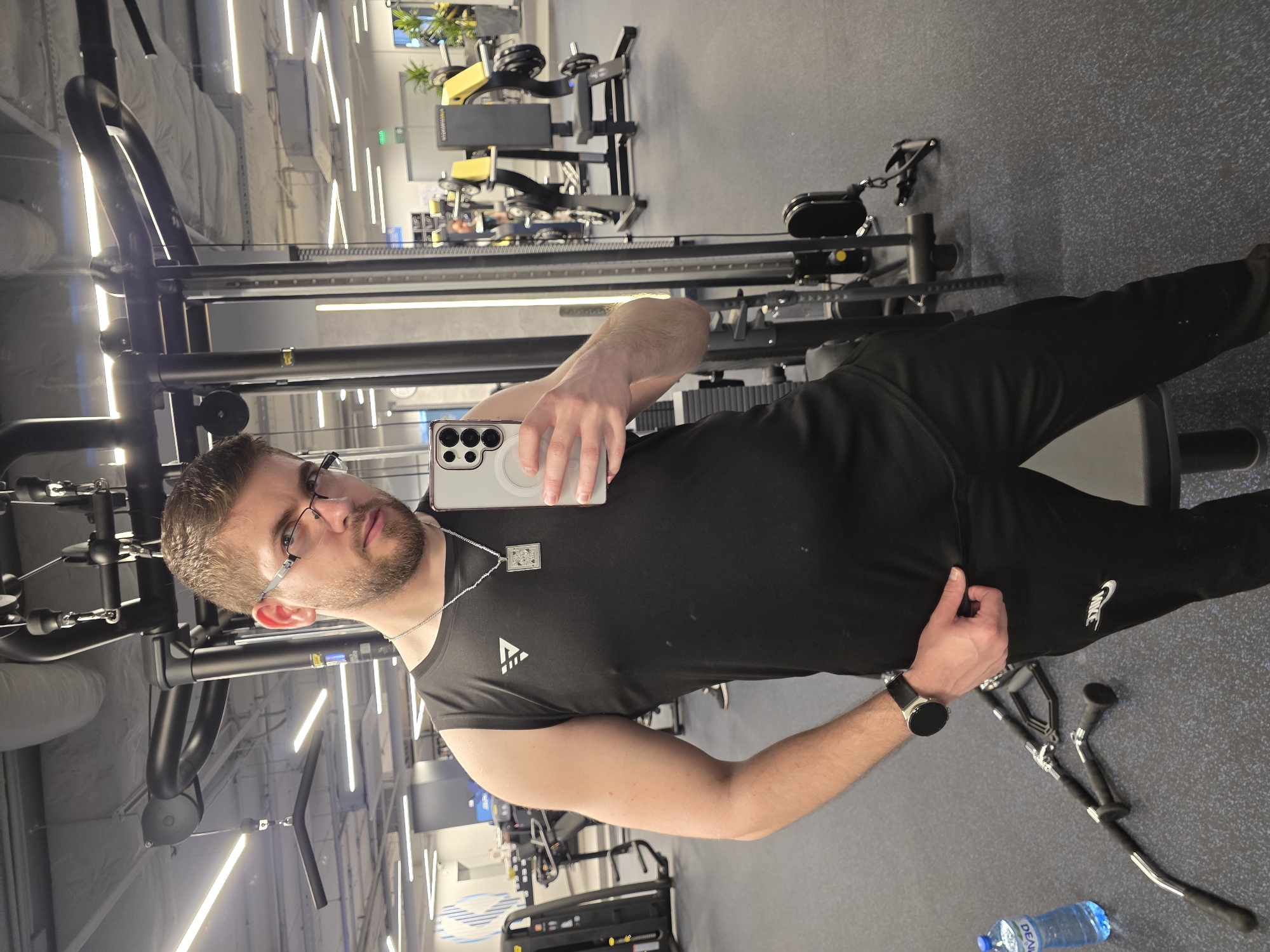 A man is in a gym with various exercise machines in the background. He is wearing glasses, a black t-shirt with a logo, black sweatpants with a white Nike logo, and a smartwatch. He is holding a smartphone to take a selfie. A pair of crutches and a bottle of water are on the floor near him. The exact location in Plovdiv, Bulgaria, cannot be determined from the image.