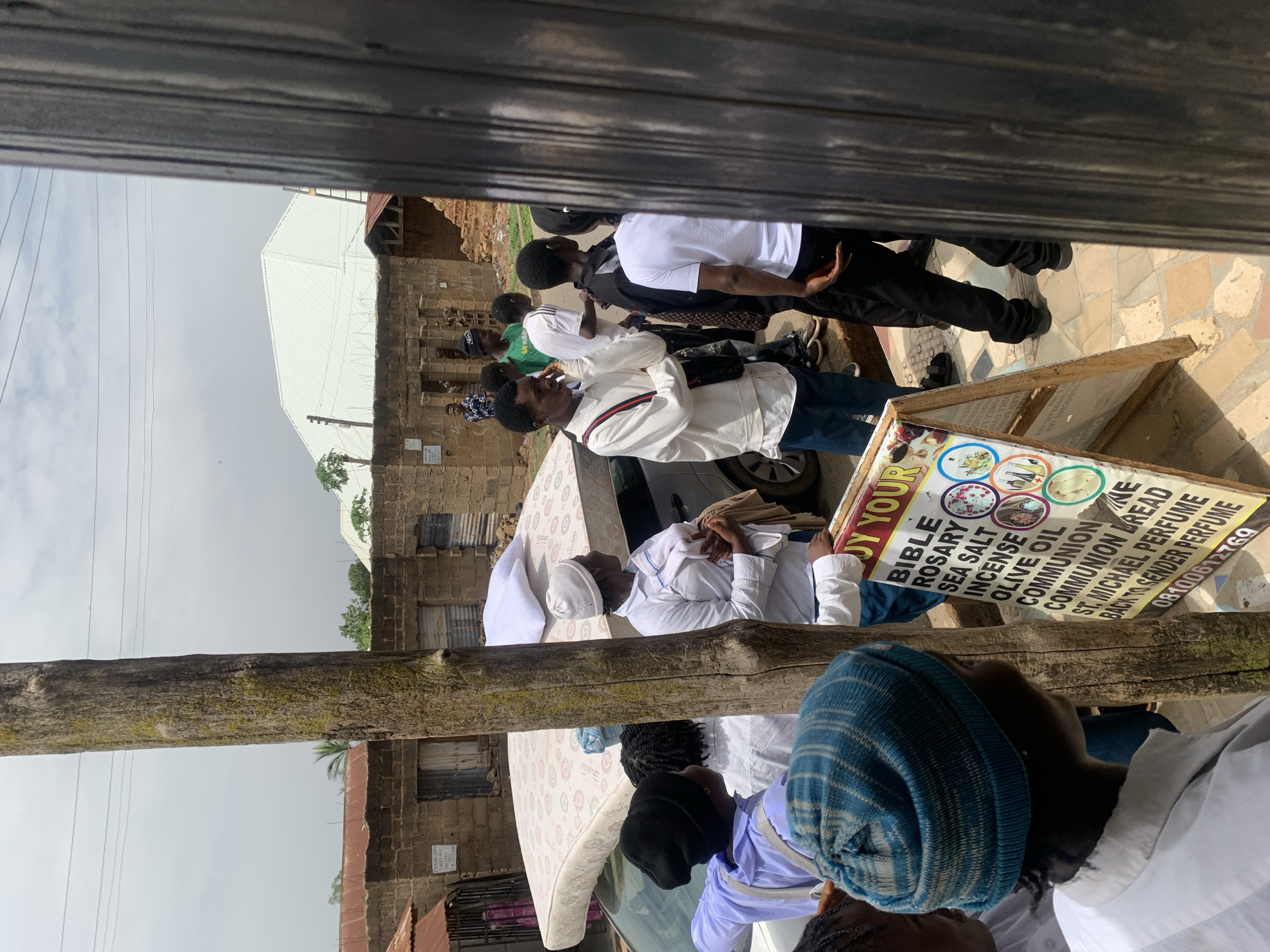 The image, taken in the city of Jos, Nigeria, captures a gathering of people, many of whom appear to be dressed in white attire, suggesting a religious or ceremonial event. Some individuals wear hats or head coverings. They are positioned around a white car. On the hood of the car is a mattress. A sign propped up nearby advertises religious items such as Bibles, rosaries, sea salt, incense, olive oil, communion bread, and St. Michael perfume. The text on the sign includes a phone number, "08100612769". The background features buildings of brick and corrugated metal, as well as a utility pole. The scene is viewed through a partial frame, likely a window or doorway.