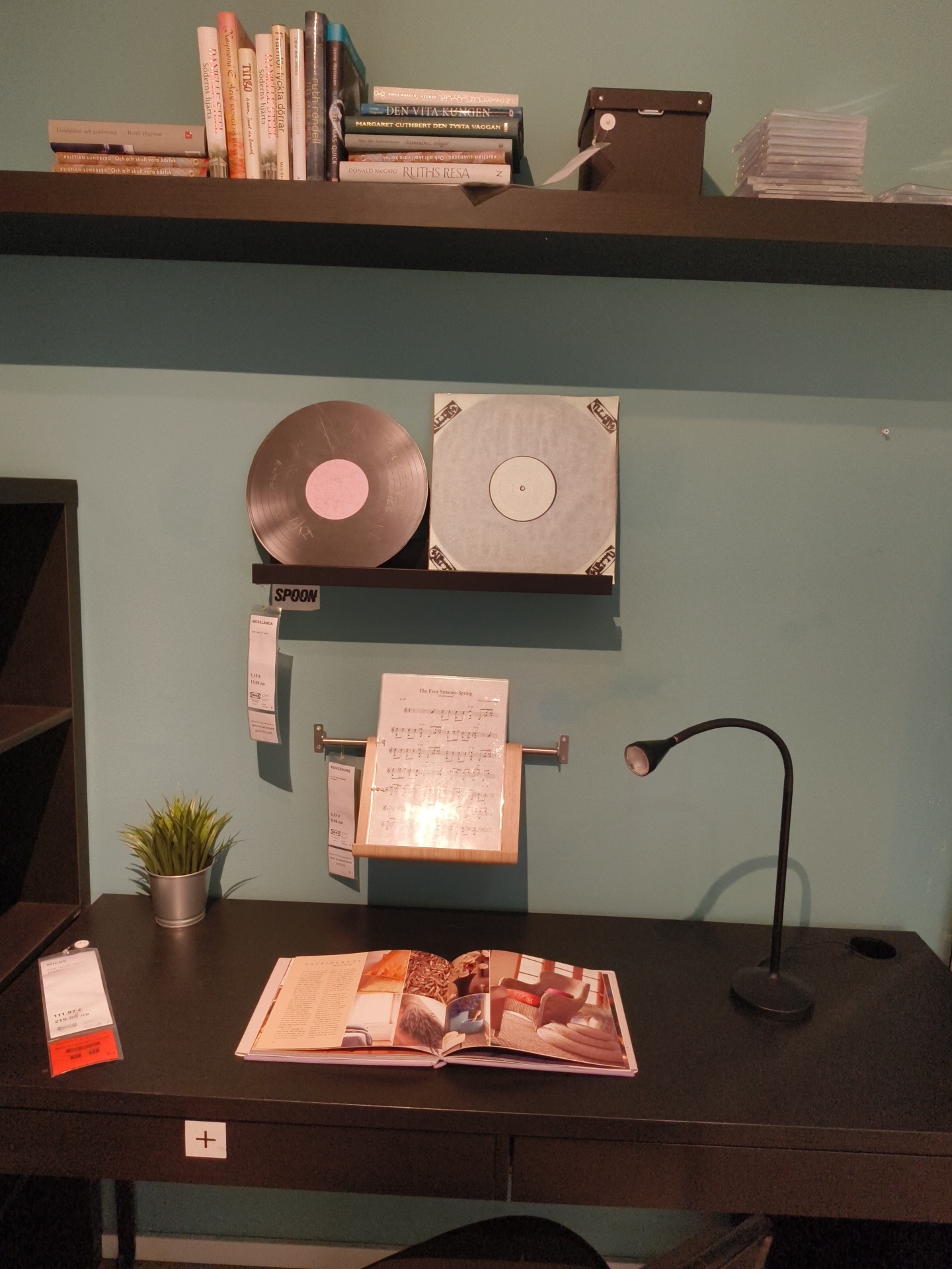 The image shows a desk setup against a light blue wall. On a shelf above the desk are books, a black box, and stacks of CDs. Two vinyl records are mounted on a smaller shelf below the main shelf. Below the records, sheet music is displayed. On the desk, there is a small potted plant, a lamp, an open catalog or book, and a price tag. The desk appears to be dark brown or black. The image was likely taken in an IKEA store, since many of the items are labeled with IKEA tags and model names. There is no indication of a location in Sofia, Bulgaria.