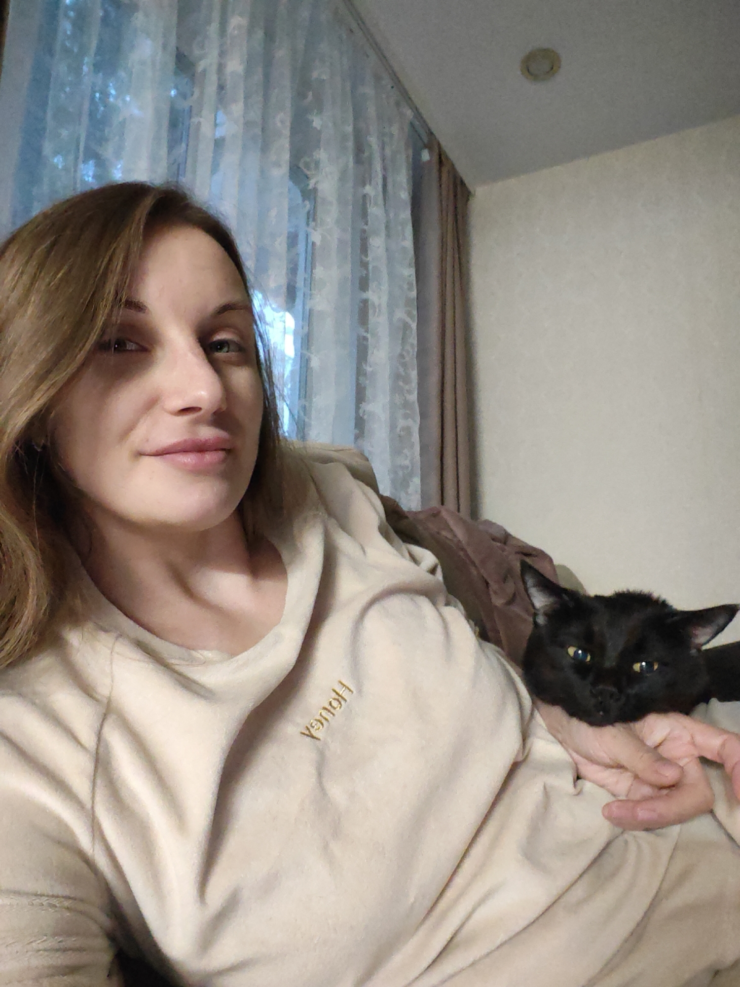 A woman is holding a black cat in her lap. The woman is wearing a light beige sweatshirt and has shoulder-length brown hair. The cat has yellow eyes and appears relaxed. They are indoors, with curtains and a wall visible in the background. The picture appears to be taken in a domestic setting and the location is Chaikovsky, Russia.