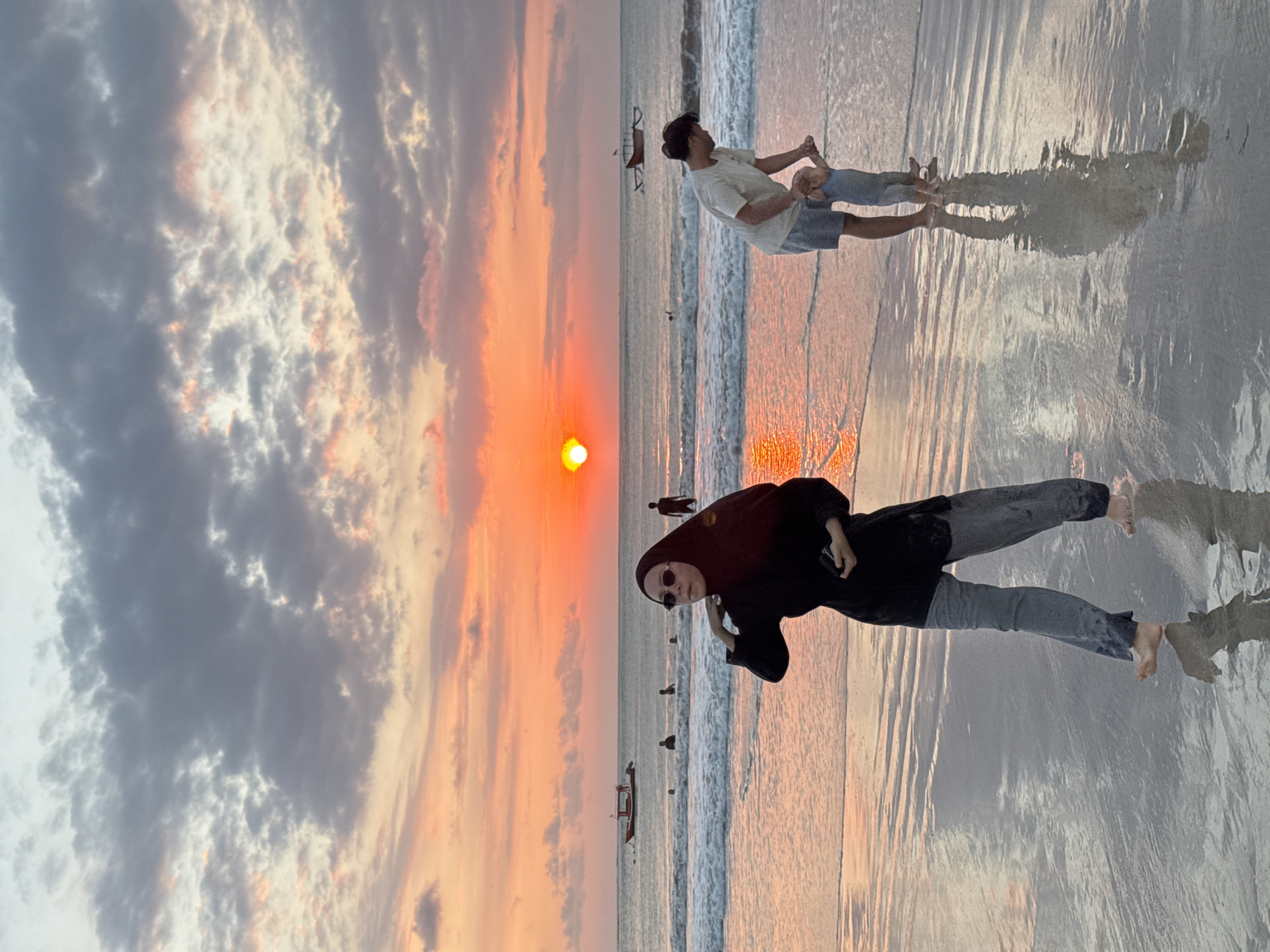 The image, taken in Kuta Selatan, Indonesia, captures a vibrant sunset beach scene. The horizon divides the frame, with the ocean and wet sand occupying the lower half and a dramatic sky filling the upper portion.
Two main subjects are prominently featured on the wet sand. On the right, a woman, identifiable by her dark hijab, dark top, light-wash jeans, and sunglasses, stands facing the camera. Her bare feet are partially submerged in the shallow, reflective water, and she appears to be posing, perhaps holding a phone or small camera in her left hand. Her shadow stretches behind her on the wet sand. On the left, further back, a man in a light-colored t-shirt and shorts holds a small child or infant in his arms, facing away from the camera towards the ocean. His bare feet are also in the water, and a long shadow extends from him. In the distance, several smaller, indistinct figures are visible on the water, along with a small, dark red boat.
The activity depicts individuals enjoying a leisure moment at the beach during sunset. The woman seems to be capturing or posing for a photo, while the man is spending time with a child by the sea.
The time of day is clearly sunset, marked by a vivid orange sun positioned on the horizon, radiating brilliant light. The sky is a spectacle of colors, with a blend of dark grey clouds and clouds illuminated with warm orange and pink hues. The calm ocean features gentle waves, and the highly reflective wet sand amplifies the beauty of the scene by mirroring the sky, the sun, and the figures. No visible text is present in the image.