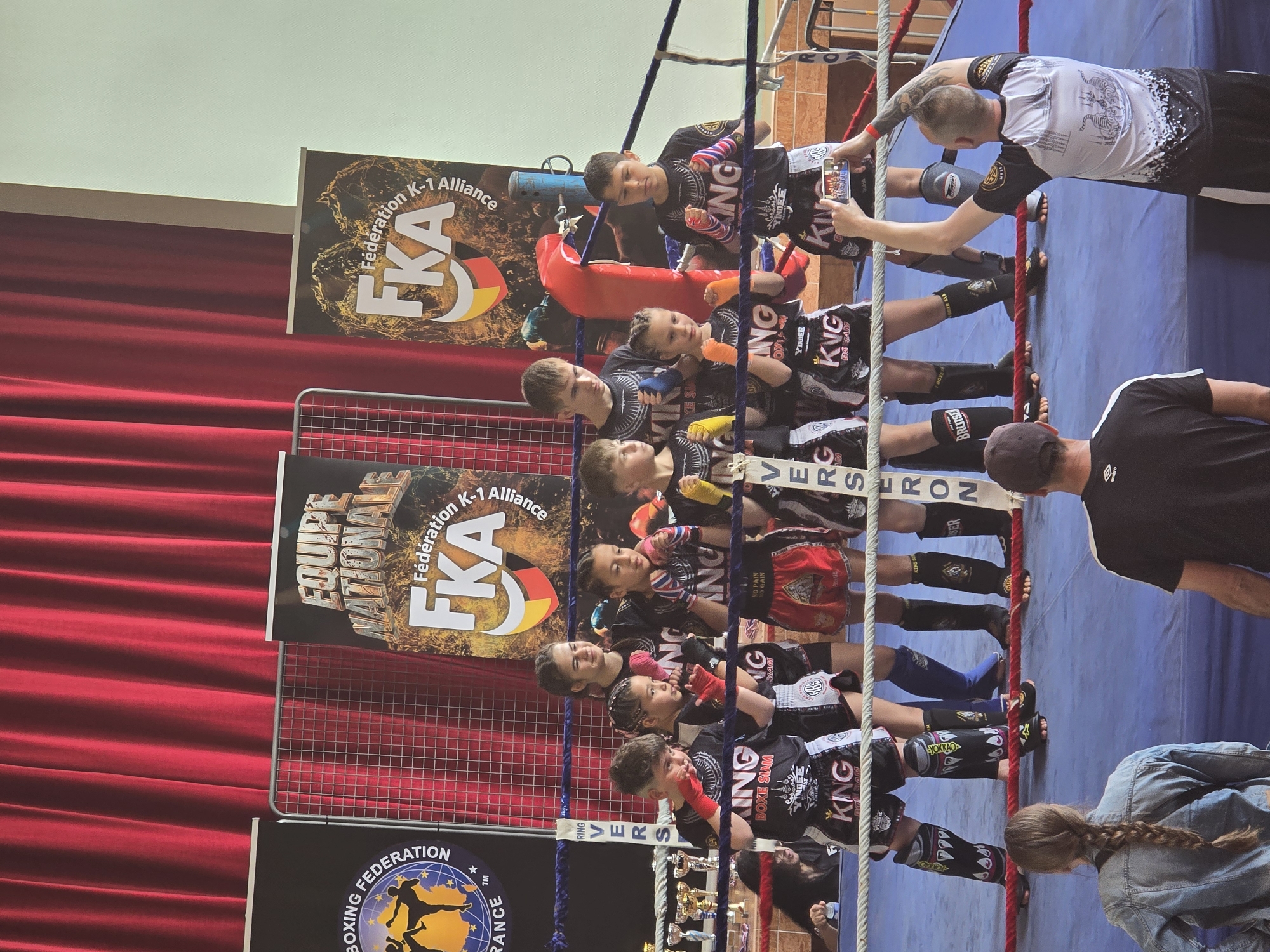 A group of young kickboxers, clad in black and gold “KING” uniforms and protective gear, line the edge of a boxing ring, likely at a competition. They are being photographed or filmed by adults standing outside the ring. Banners with the logos "Fédération K-1 Alliance," "EQUIPE NATIONALE," and "BOXING FEDERATION FRANCE" adorn the background, along with a red curtain. Trophies are visible in the lower center of the image. The location is Berlaimont, France, based on the text on the banners.