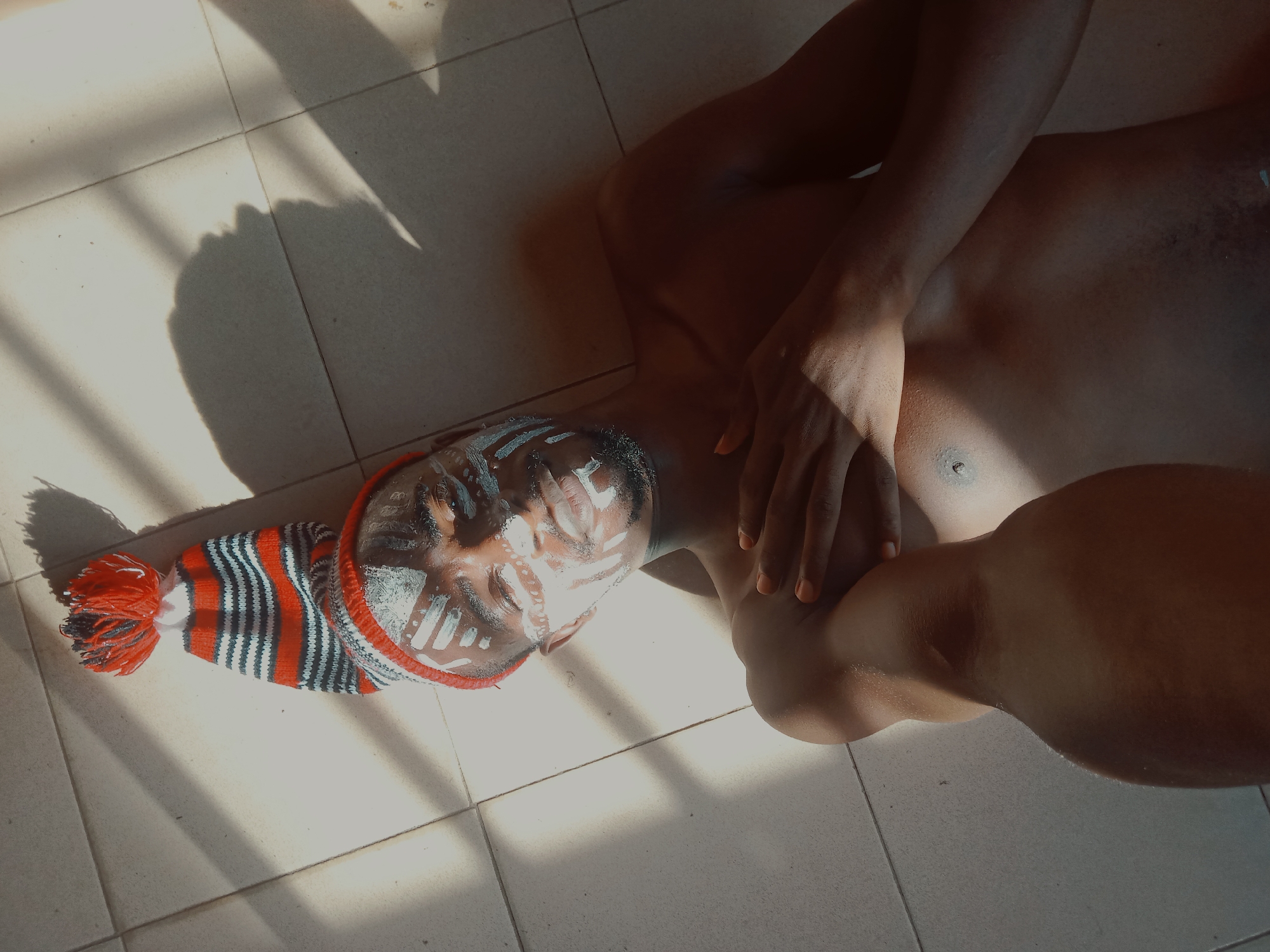 A brown-skinned individual with tribal white markings on their face and wearing a red, black, and white striped hat is lying on a tiled floor. The person's torso is partially visible, with one arm across the chest. Sunlight casts shadows across the floor and the person's body. The location is in the city of Unknown, Unknown.