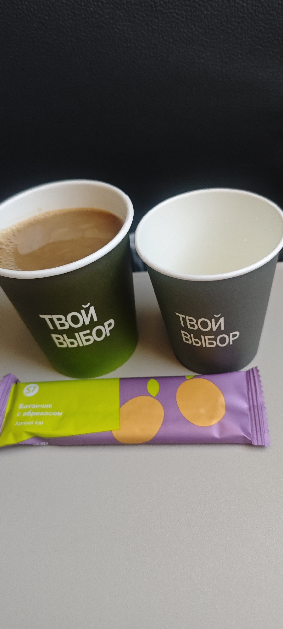 On a light gray surface, likely a tray table, are two paper cups and a wrapped apricot bar. The cup on the left is filled with a light brown liquid, presumably coffee or tea with milk, while the cup on the right is empty. Both cups are dark green with the white text "ТВОЙ ВЫБОР" printed on them. Below the cups is a light purple and green bar labeled "Apricot Bar" in both Russian and English. The image was likely taken in an airplane cabin, as suggested by the dark black headrest visible in the background. The text on the cup, "ТВОЙ ВЫБОР" translates to "Your Choice." While "Your Choice" is a common advertising phrase and does not immediately imply that this is from Russia, "Батончик с абрикосом", which translates to "bar with apricot", written in Russian on the apricot bar indicates the location is most likely in Russia. Further, the apron bar is manufactured by a company in Novosibirsk, Russia.