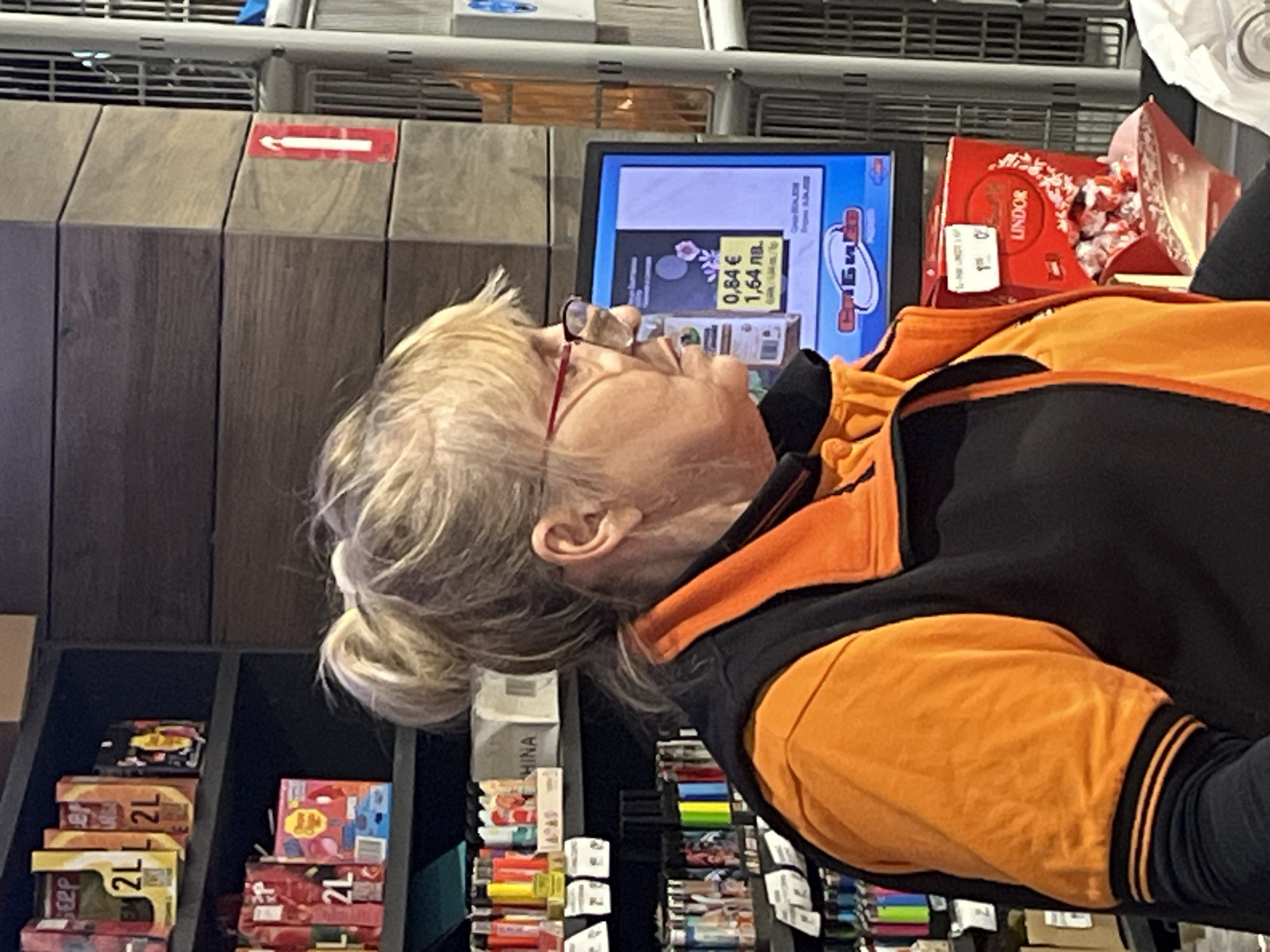 A middle-aged woman with blonde hair pulled into a low bun is looking at a digital display that shows the price of 0.84€ / 1.64 лв. A red box of Lindt Lindor chocolates is to the right of the display. The woman is wearing glasses with red frames and a black and orange jacket with the sleeve cuff visible. The background shows shelves filled with boxes of various items, suggesting she is at a checkout or store. A red sign with a white arrow is visible above the shelves. Based on the price in Bulgarian levs (лв), the location is Stara Zagora, Bulgaria.