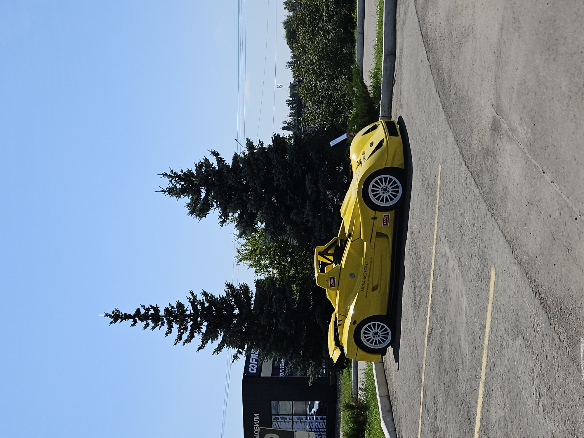 A bright yellow, open-top sports car is parked in an outdoor parking lot. The car has white rims and visible branding, including "LADA" logos. Behind the car, a dark building with signage that includes "CO FRESH" and Russian text for "MOBILES" is visible. Two tall evergreen trees stand behind the building. The parking lot is asphalt, marked with yellow lines. The background consists of green bushes and trees under a clear blue sky. The location is identified as Ulyanovsk, Russia, at 20 Yuzhnyy Pyl, 67.