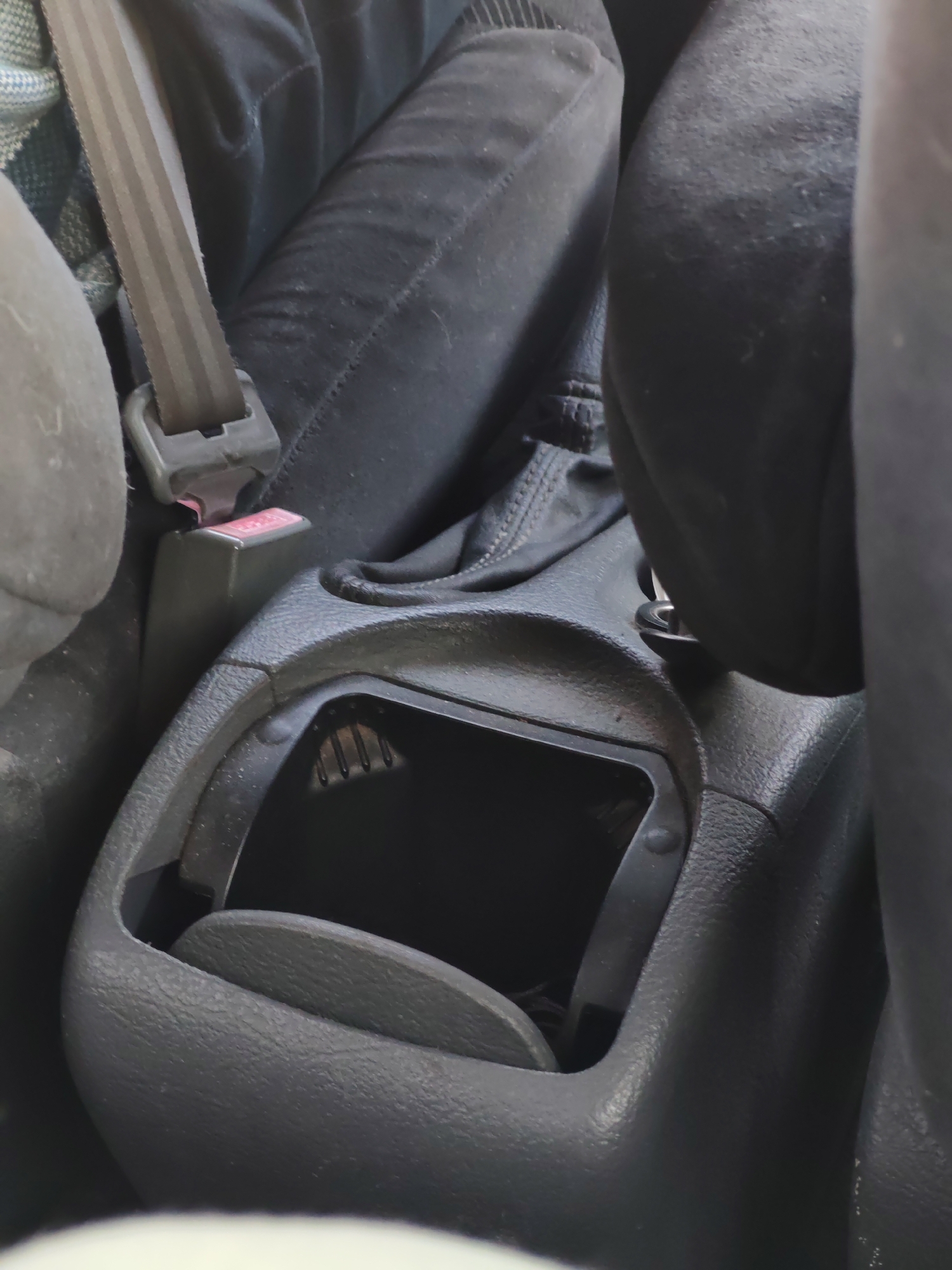 The image shows the interior of a vehicle. A black plastic center console is prominent, with a partially opened storage compartment revealing a dark interior. A gear stick with a black leather boot is visible nearby. To the left, a seatbelt buckle is fastened. The surfaces appear dusty. The location is Unknown, Unknown.
