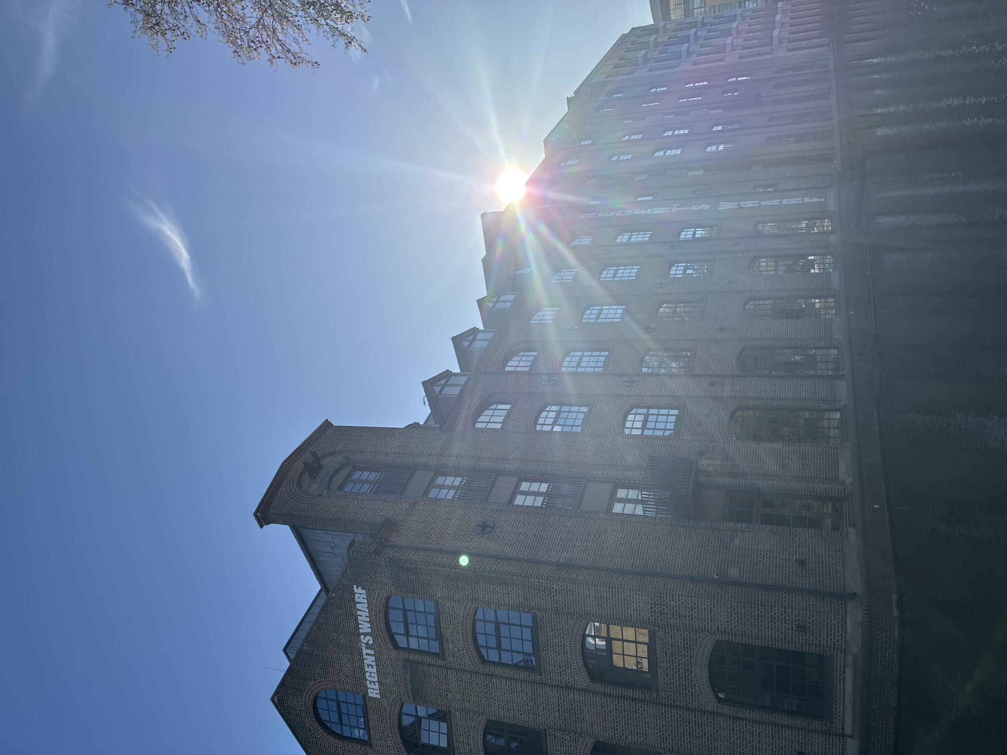 The image shows a low-angle view of Regent's Wharf in London, United Kingdom, a multi-story brick building with numerous windows and rounded arches. The building is situated under a bright blue sky with a few wispy clouds. The sun is positioned behind the building, creating a lens flare effect. Part of the sky and a leafy tree branch can be seen in the upper left corner of the frame. A dark wall with windows is visible to the right.