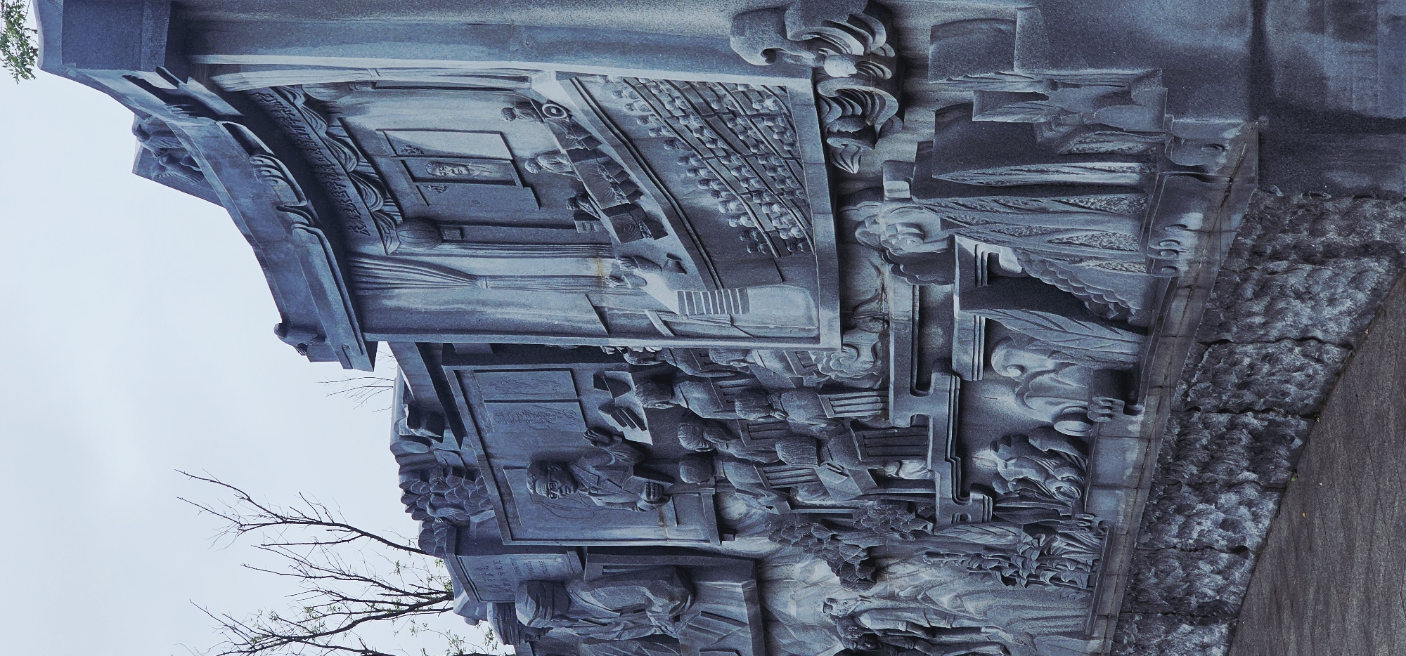 This image shows a detailed bas-relief sculpture located in the Chongqing High-tech Zone, China. The sculpture depicts a scene with multiple figures, likely representing an educational or historical event. In the foreground, a group of individuals are seated, appearing to be students or an audience, facing a central figure who stands and gestures. This central figure is dressed in formal attire and holds a book. Behind him, a framed portrait is visible. The sculpture extends to the right, showing more rows of seated figures and architectural elements such as draped curtains. To the left, other figures and decorative motifs are carved into the stone. The sculpture is rendered in a monochromatic, grey stone, and is set against a backdrop of a pale sky with some bare tree branches visible. The base of the sculpture is made of rough-hewn stone blocks.