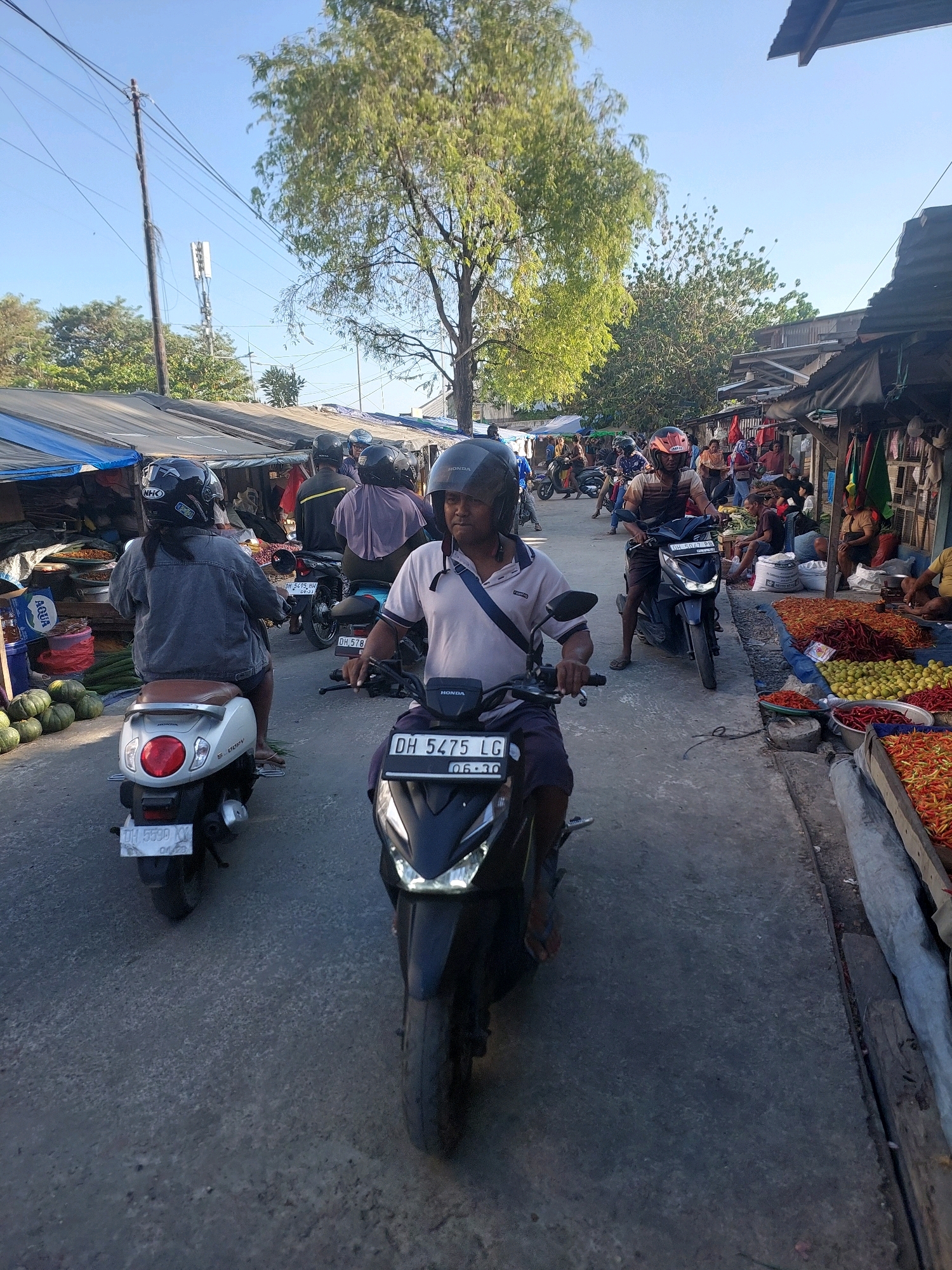 This is a street-level, eye-level, medium shot of a busy marketplace scene in Kupang, Indonesia. The foreground is dominated by a man riding a black Honda scooter, wearing a light-colored polo shirt, dark shorts, and a black helmet. He is looking forward. Behind him, a woman on a white Honda scooter, wearing a denim jacket and dark shorts, also with a helmet, is visible. Other motorcyclists and pedestrians are further down the street, navigating the crowded market stalls that line the right side of the road. These stalls display a variety of produce, including chilies and lemons. The environment appears to be a sunlit urban area with a clear blue sky. A cell tower and trees are visible in the background. The overall impression is one of a lively and active marketplace.