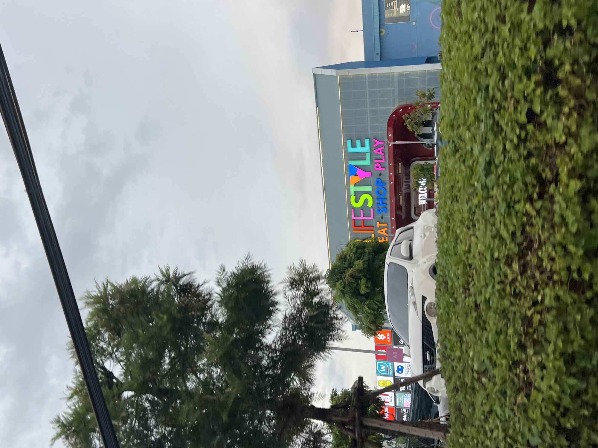The image shows a white car parked in front of a building with "LIFESTYLE EAT SHOP PLAY" written in colorful letters. The building appears to be a mall or a commercial complex. There are trees and bushes in the foreground and background. The sky is cloudy. The scene is captured outdoors, likely on a street or parking lot. The presence of commercial signage suggests an urban or suburban setting. The word "Chalong" is not visible in the image.