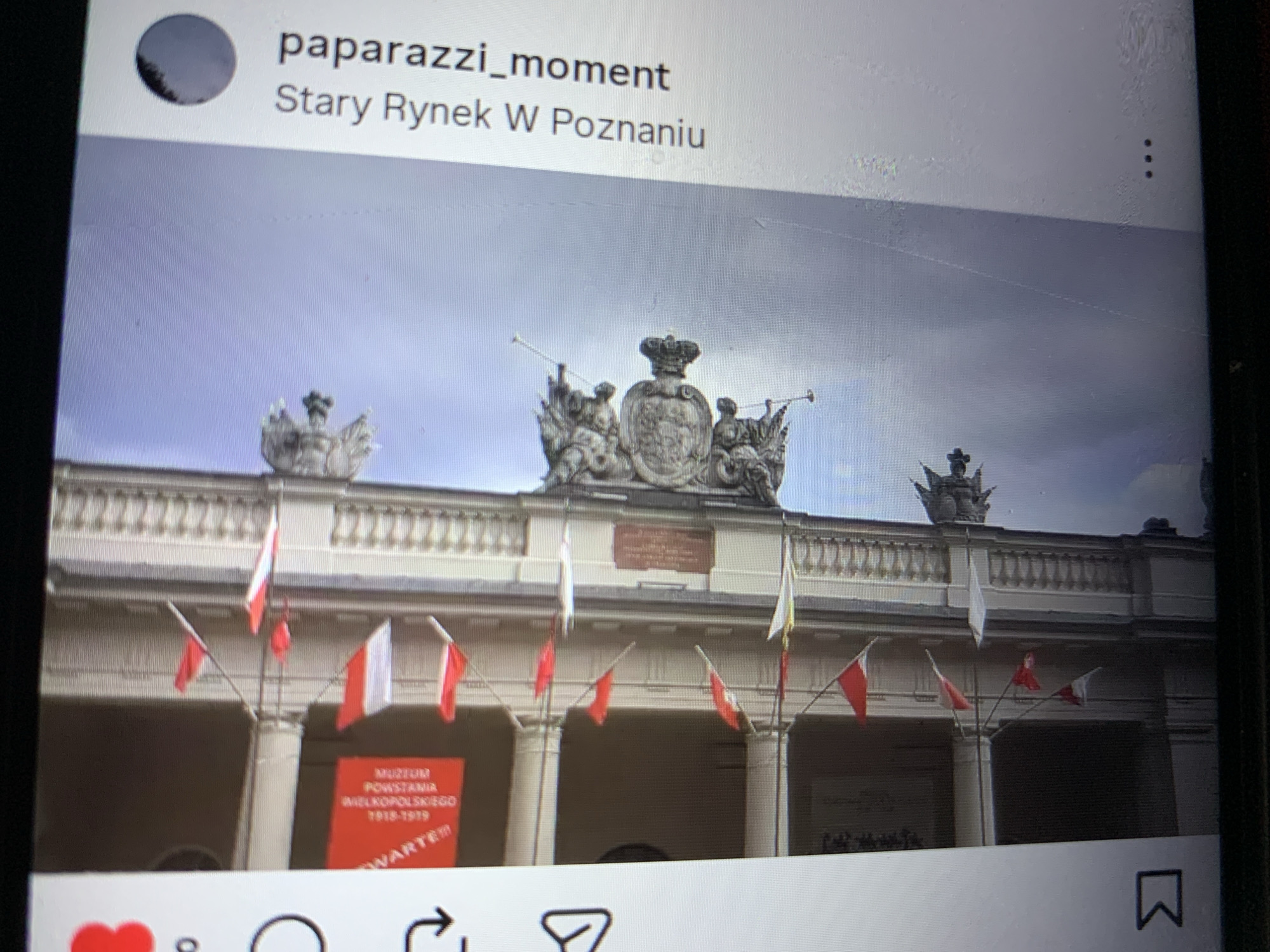 The image is a photograph taken from a screen, likely a mobile device, showing a building with architectural details and flags. The flags are red and white, and appear to be Polish flags, suggesting the location may be in Poland. The building has a colonnaded facade and decorative elements, including statues and crests. The sky is overcast. Text is visible on the right side of the screen, reading "paparazzi_moment" and "Stary Rynek W Poznaniu", which translates to "Old Market Square in Poznań". There are also some interface elements visible on the left side, indicating it is a screenshot from an application. The image does not show anything related to Lymanka, Ukraine.