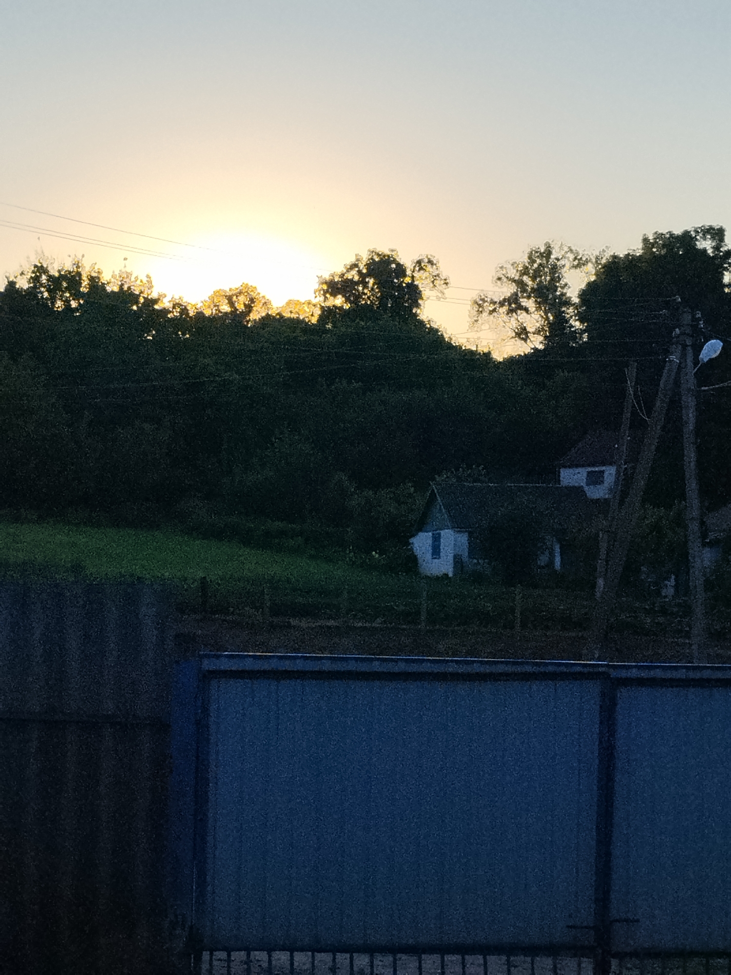The image depicts a rural scene at dusk, likely in Ustyvytsia, Ukraine, with the sun setting behind a dense line of trees. In the foreground, a dark blue corrugated metal gate dominates the bottom portion of the frame. Behind the gate, a dark, rough-textured wall is visible on the left. The middle ground features a sloping hill covered with dark green trees. Nestled within the trees are two white houses with dark roofs. A wooden utility pole with a streetlight is present to the right of the houses. The sky is a pale gradient of yellow and blue, indicating the setting sun. The overall impression is one of a quiet, twilight evening in a rural setting.