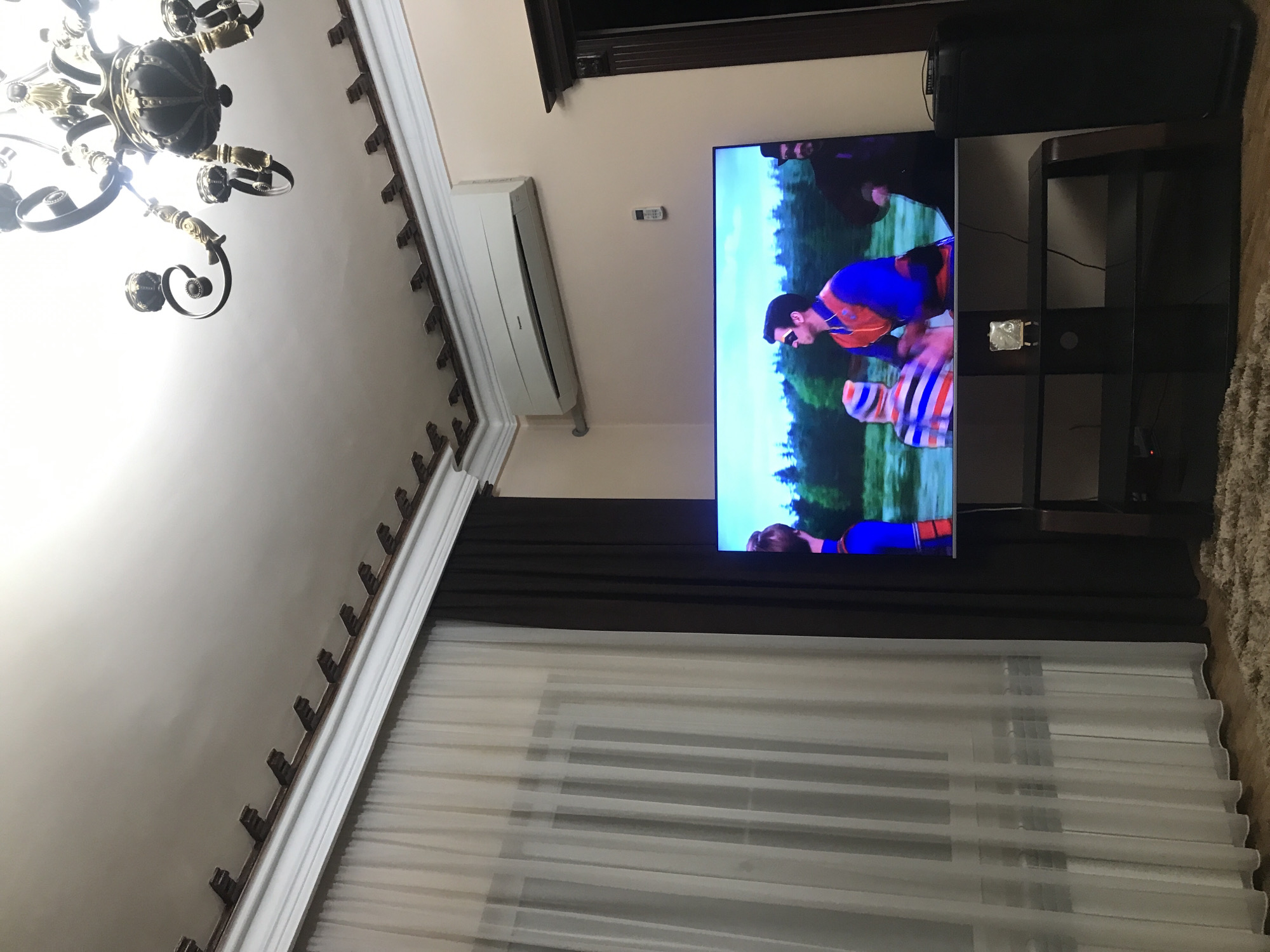 A residential interior in Yerevan, Armenia, featuring a ceiling chandelier, white ceiling, cornice molding, wall-mounted air conditioning unit, curtains, and a flat-screen television displaying an image of people in costume. The television is mounted on a dark-colored stand. Adjacent to the television stand is a speaker and a rug. The window is covered by sheer white curtains.