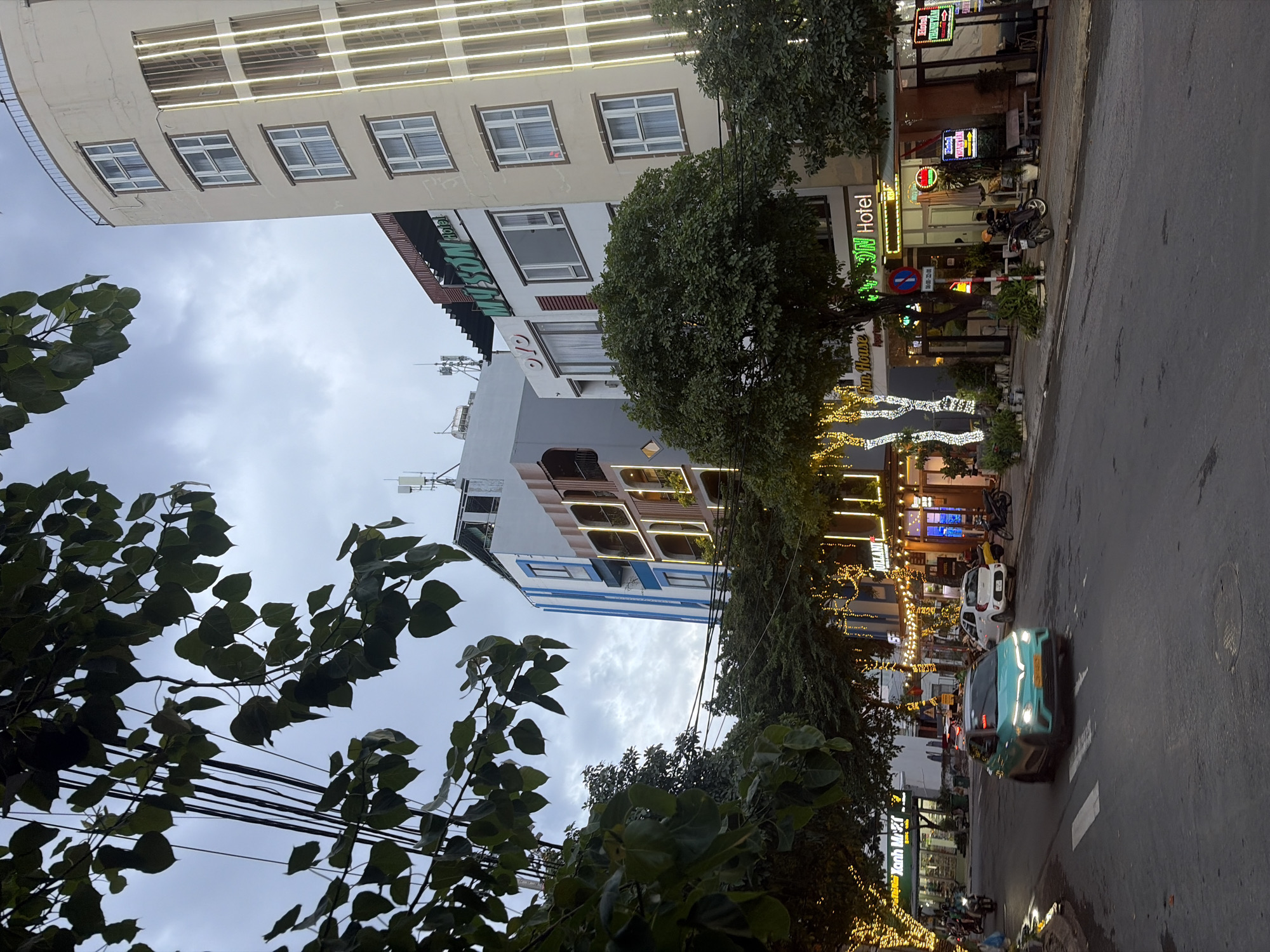 The image depicts a street scene in the city of Đà Nẵng, Vietnam, at dusk or early evening, as indicated by the artificial lighting from buildings and street decorations. In the foreground, a teal-colored electric car is driving down the road, crossing a pedestrian walkway. On the right side of the street, there are several multi-story buildings with businesses on the ground floor, including a hotel sign that reads "MY SON HOTEL". The buildings are adorned with string lights, creating a festive atmosphere. Trees with lush green foliage are planted along the sidewalks and street, some of which are also decorated with lights. Several motorcycles and scooters are parked along the sidewalk, and some are visible in motion on the street. Overhead, a dense network of electrical wires crisscrosses the sky, a common sight in Vietnamese cities. The sky is overcast with a cloudy, grayish hue, suggesting an impending change in weather. The overall scene conveys a sense of urban activity and commerce in a Southeast Asian setting.