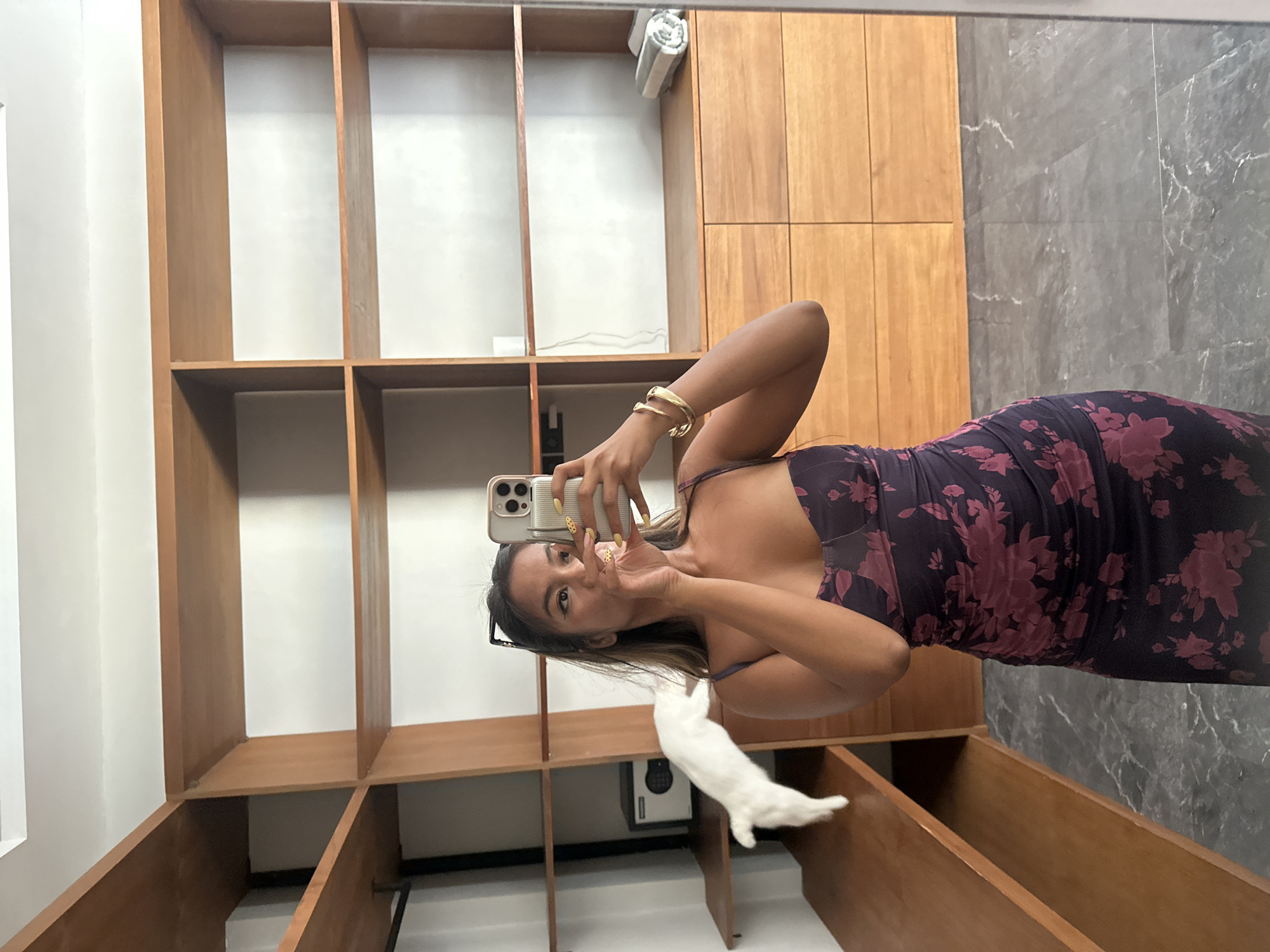 The media file is a vertical, medium-shot, mirror selfie taken by a young woman. She is wearing a dark purple, floral-patterned dress. She holds a smartphone in her right hand, capturing her reflection. A gold bangle adorns her left wrist. She is standing in front of a large, wooden shelving unit that appears to be part of a closet or dressing room. To the right of the shelves, there are wooden cabinets and a grey marble wall. A white, fuzzy object, possibly a stuffed animal or a garment, is visible on the shelves below her. The setting suggests an indoor location in North Kuta, Indonesia, likely a home or boutique. The lighting appears to be artificial, suggesting it could be any time of day. The woman's facial expression is playful, with pursed lips and wide eyes looking into the camera. There is no visible text in the media file.