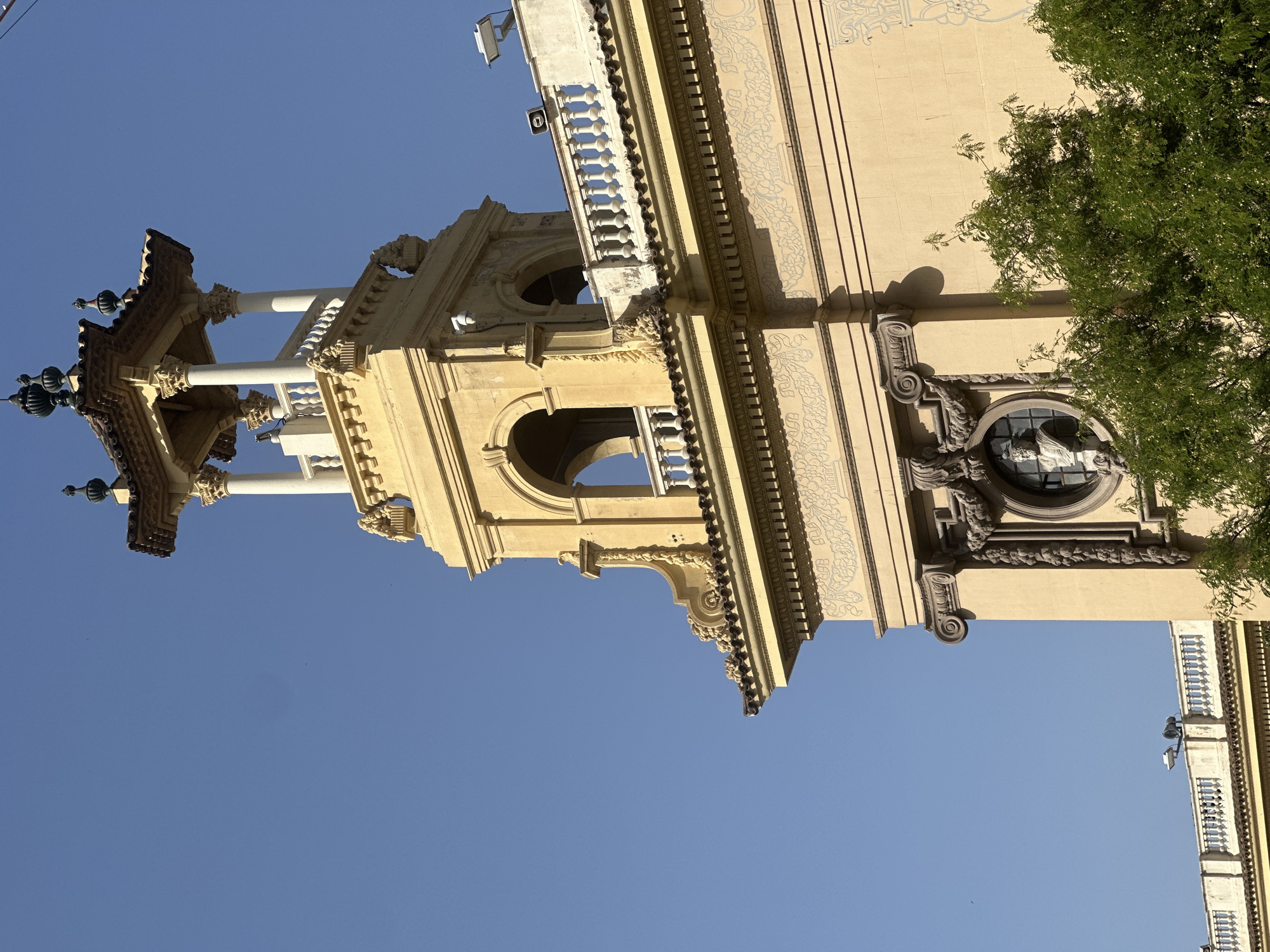 The image shows a close-up of a yellow building with ornate architectural details. A tower-like structure with a tiled roof, supported by white columns, rises from the building. The building's facade features arched openings, a decorative railing, and intricate carvings around a circular window containing a bust. Green foliage from a tree partially obscures the lower portion of the building. The sky above is clear and blue. This building is likely located in Barcelona, Spain, and appears to be a historical or landmark structure.