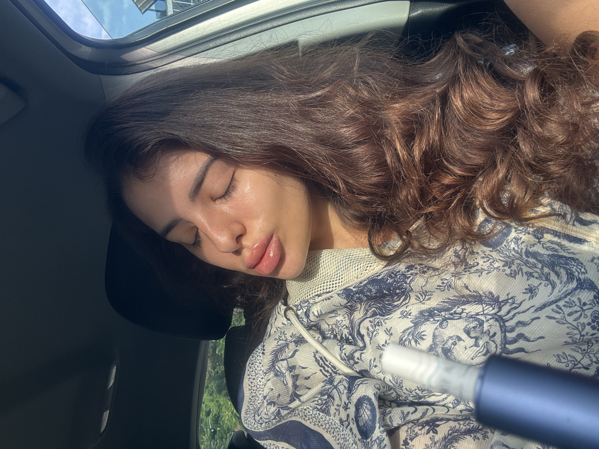 In the close-up image, a person with long, dark brown hair is seen inside a vehicle. The person's eyes are closed, and their lips are slightly puckered, suggesting a pose or a moment of relaxation. Sunlight illuminates the person's face, highlighting their features. They are wearing a white and blue patterned garment, possibly a jacket or hoodie, with a white drawstring. In the foreground, a portion of a blue cylindrical object, possibly a vape pen or a similar device, is visible. The background shows the interior of the car, including a headrest and part of a window with a glimpse of greenery and a blue sky outside. There is no specific indication of the location within the city of Plovdiv, Bulgaria.