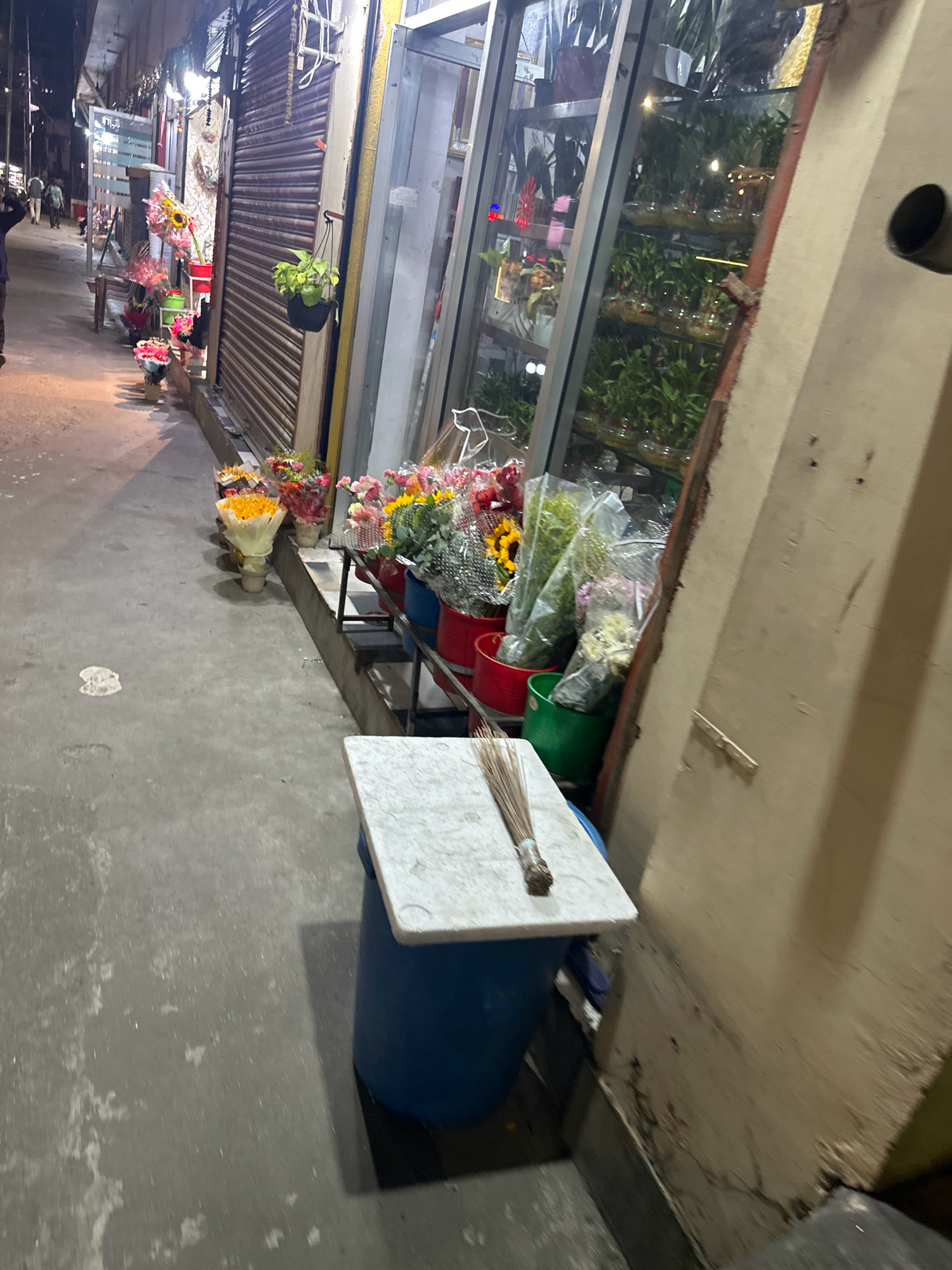 The image depicts a sidewalk flower stall on a street in Kathmandu Metropolitan City, Nepal. Several bouquets of flowers, including sunflowers and other colorful blooms, are displayed in buckets and vases along the curb. In the background, a storefront with rolling metal shutters is visible. A few potted plants hang from the store's facade. A broom rests on a blue barrel topped with a white marble slab in the foreground. The street is paved with concrete, and some litter is scattered on the ground. The overall scene suggests a busy urban environment with a focus on commerce and street-side vendors.