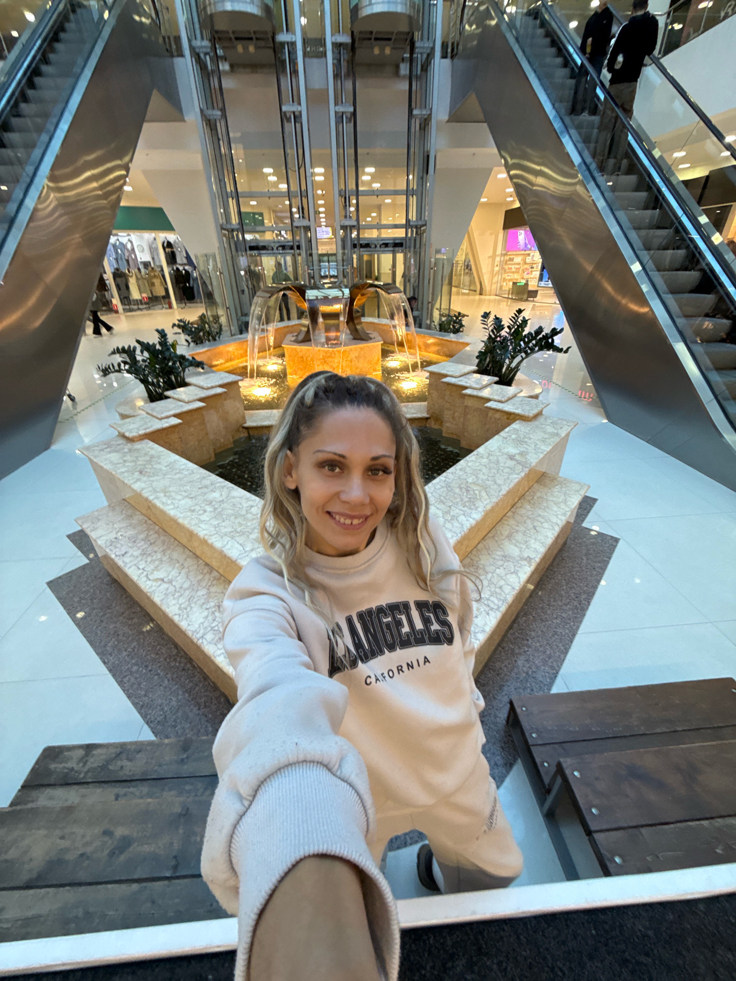 This is a selfie of a woman in a shopping mall. She is wearing a light-colored sweatshirt with "LOS ANGELES CALIFORNIA" printed on it. She has blonde hair styled in a ponytail and is smiling at the camera. In the background, there is a multi-tiered fountain with water flowing. Escalators and an elevator shaft are visible on either side of the fountain. The floor is tiled, and there are some wooden benches in the foreground. Shops and other people are visible in the distance. The location of Rusanovo, Russia is not depicted in the image.