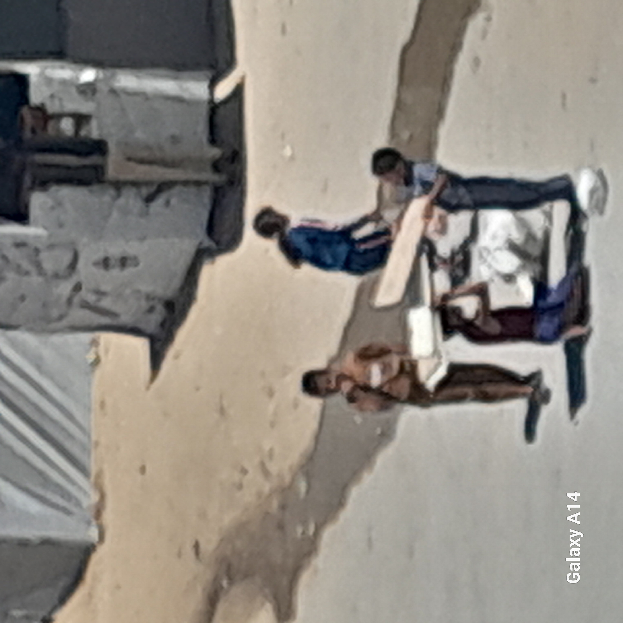 Four individuals, appearing to be children, are gathered around some flat, rectangular objects on a dirt and paved surface. One individual, wearing a brown shirt, stands to the left, looking down. Another individual, wearing a blue and white striped top and dark shorts, is seated on a wooden plank to the right. A third individual is crouched down, appearing to carry a plank on their head. A fourth individual, dressed in a blue shirt and dark pants, stands to the right, also interacting with the planks. The scene is set outdoors, with a building or structure visible in the background. The ground is a mix of light-colored dirt and darker pavement. The overall impression is of children engaged in activity with wooden planks in an outdoor, possibly urban or semi-urban environment.