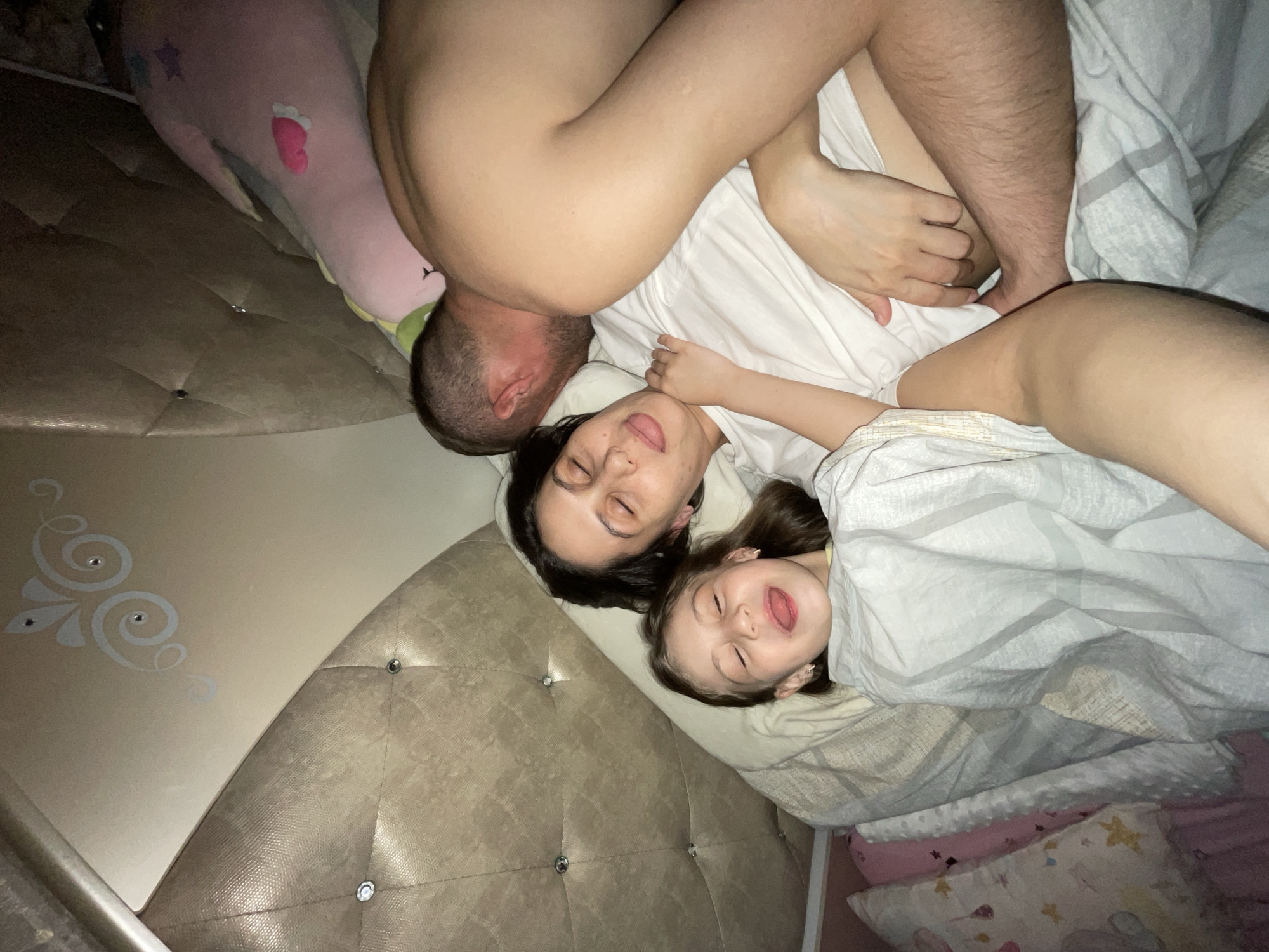 A man, woman, and child are lying in a bed. The man is positioned with his back towards the camera and his head near the headboard. The woman is lying next to him, eyes closed. A child is lying next to the woman, also with eyes closed and tongue sticking out. They are covered with blankets. A pink stuffed animal is near the headboard. The location is unknown.