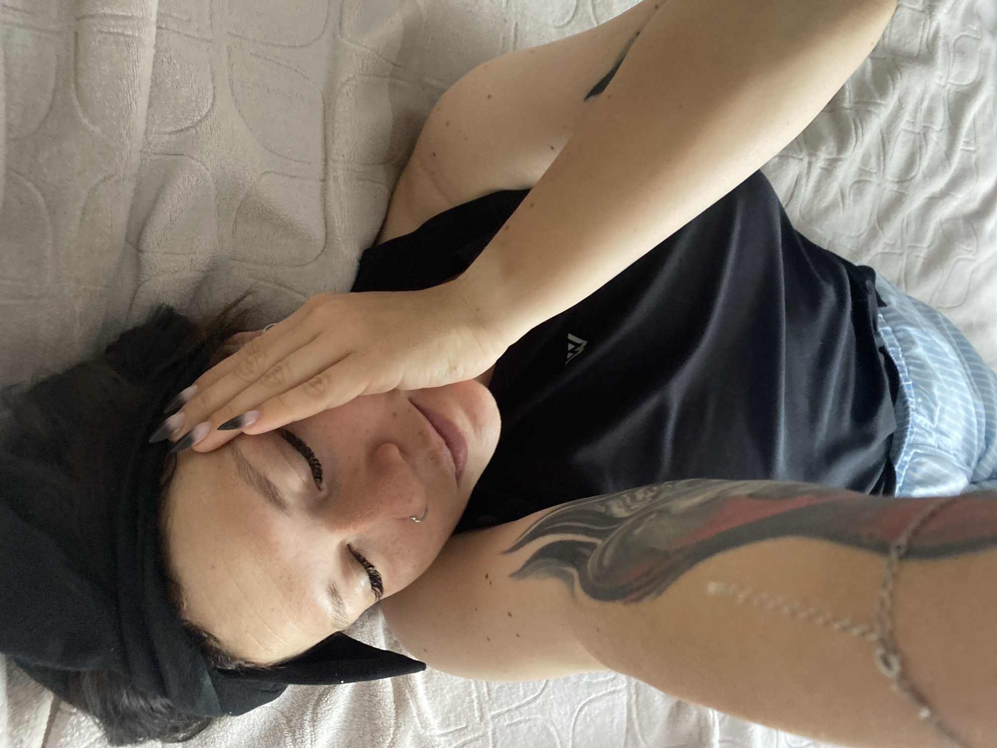 An overhead shot shows a person lying on their back on a bed. The person is wearing a black tank top and blue and white striped shorts. Their right arm is bent and resting on their forehead, covering their eyes. Their left arm is raised and bent, with a silver chain bracelet on their wrist. The person has a nose ring and tattoos on their upper left arm. The bed has a textured light grey blanket. The scene appears to be indoors.