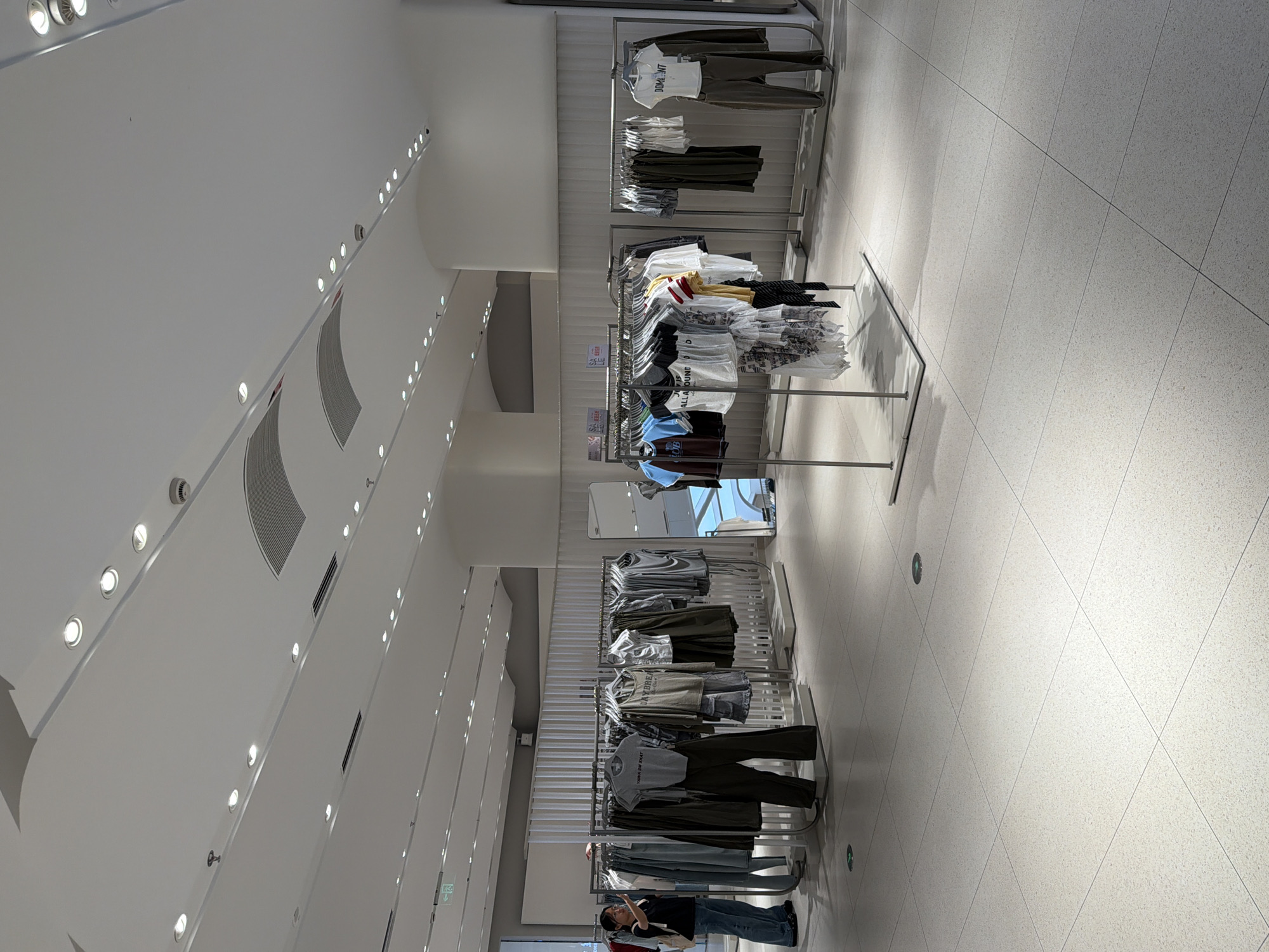 The image depicts the interior of a clothing store in Qinhuai District, China. The store features a modern design with white tiled floors and a white ceiling equipped with numerous recessed lights and ventilation grilles. Clothing racks with various apparel, including t-shirts and pants, are displayed throughout the store. A woman wearing a black top, blue jeans, and carrying a white backpack stands on the left, looking at her phone. She is positioned near a clothing rack displaying several pairs of pants and shirts. In the mid-ground, several clothing racks hold a variety of tops and pants, some with price tags visible. To the right, a large rectangular mirror is propped against a wall. Another clothing rack with t-shirts and pants is visible further to the right. The overall environment is clean, well-lit, and organized, typical of a retail clothing establishment.
