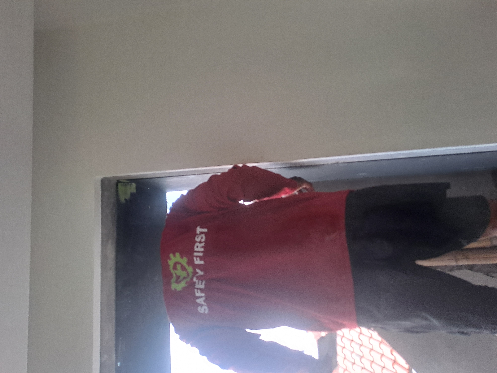 A person, viewed from behind, stands in a doorway. The person is wearing a long-sleeved red shirt with the words "SAFETY FIRST" printed in white letters on the back, and a green logo above the text. The person is also wearing dark shorts. The person appears to be indoors, with a bright light source visible through the doorway, suggesting it is daytime and the person is facing outwards. The environment appears to be under construction or renovation, as suggested by the unfinished doorway and wall. The person is standing near a roof with terracotta tiles, which may indicate a residential or commercial building. This scene is located in Ngaglik, Indonesia.