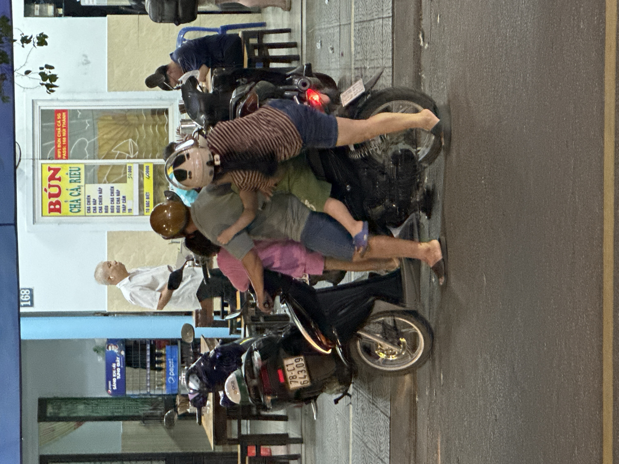 In Đà Nẵng, Vietnam, at street level, four people are crowded onto a black scooter parked on the side of a road. The scooter is facing right and is slightly angled towards the viewer. The people are: a man in the front, wearing a white short-sleeved shirt and dark shorts, with white earbuds in his ears and a light-colored helmet. Next to him, behind the man and slightly to the right, is a person in a pink shirt and shorts, also wearing a helmet. To the right of the man in front, and on the scooter, is a person in a striped brown and white long-sleeved shirt and blue shorts, with a light-colored helmet. The fourth person, in a gray shirt and blue shorts, is positioned behind the person in the pink shirt and is also wearing a helmet. The person in the striped shirt appears to be holding onto the person in the gray shirt. The scooter's license plate reads "78-C1 643.09".
In the background, there is a building with a sign that says "BÚN CHẢ CÁ, RIÊU". The sign also lists prices and the Wi-Fi password. To the left of the scooter, tables and chairs are visible, suggesting an outdoor seating area for a restaurant. The overall scene suggests a busy urban environment, likely in the evening, given the lighting conditions.