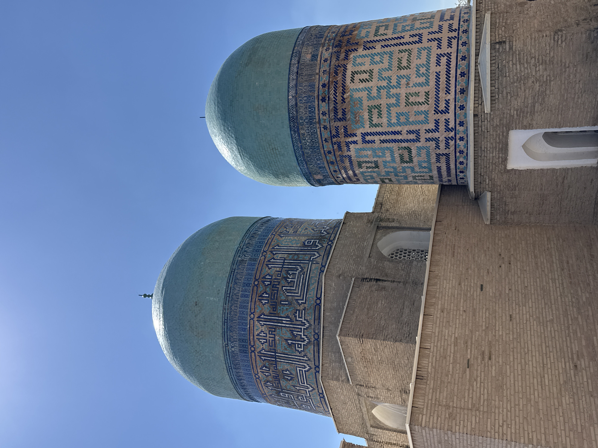 The image shows the turquoise domes of the Kukeldash Madrasah, an Islamic educational institution, in Samarkand, Uzbekistan. The domes are adorned with intricate blue and white geometric tile patterns. The structures are made of tan brick. The sky is clear and blue. The sun is shining.