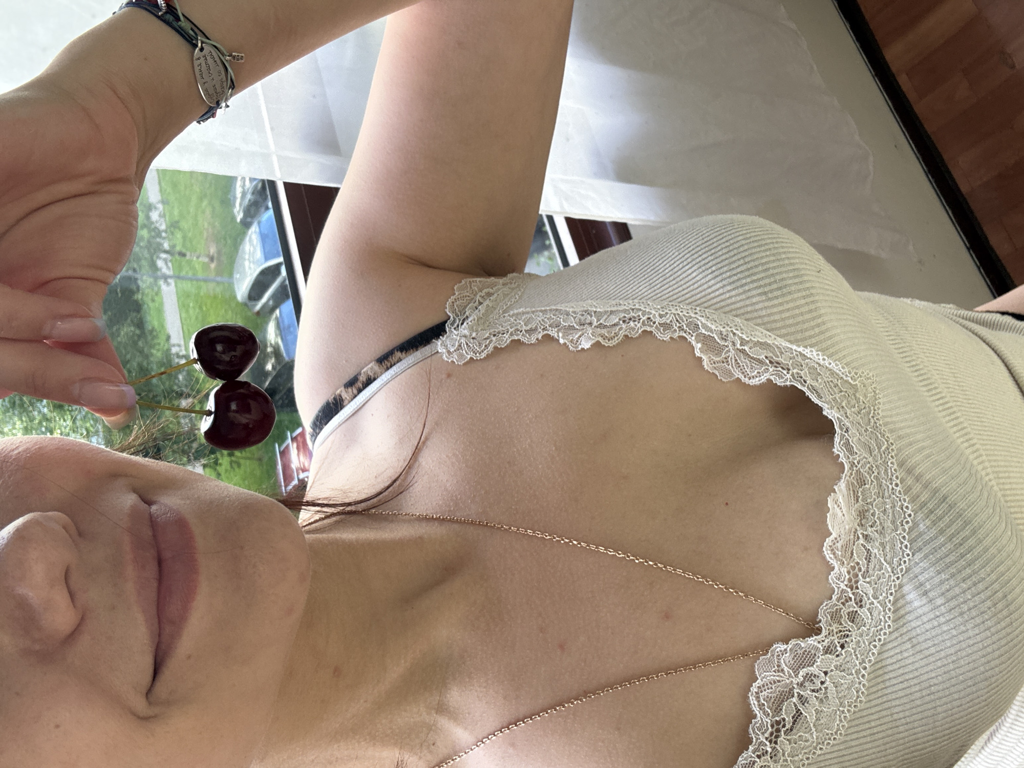 A woman is holding two cherries by their stems near her mouth. She is wearing a white ribbed tank top with lace trim. A delicate gold chain necklace is visible around her neck. On her wrist, she wears a woven bracelet with a small silver charm. The background appears to be a window with a view of trees and cars outside.