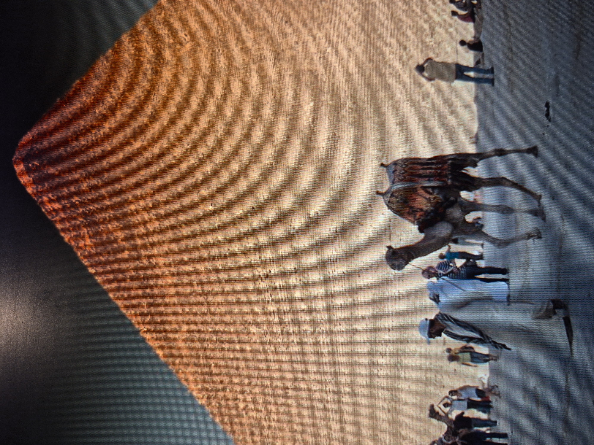 The image depicts a portion of a pyramid, likely in Egypt. To the right of the pyramid, on a sandy surface, there is a camel with a decorated saddle and a group of people. The people appear to be tourists or locals, some wearing traditional clothing. Some are walking near the camel. The image is rotated, so the scene appears sideways. It shows a tourist scene with a camel in front of a pyramid. There is no evidence to suggest it is in Yerevan, Armenia.