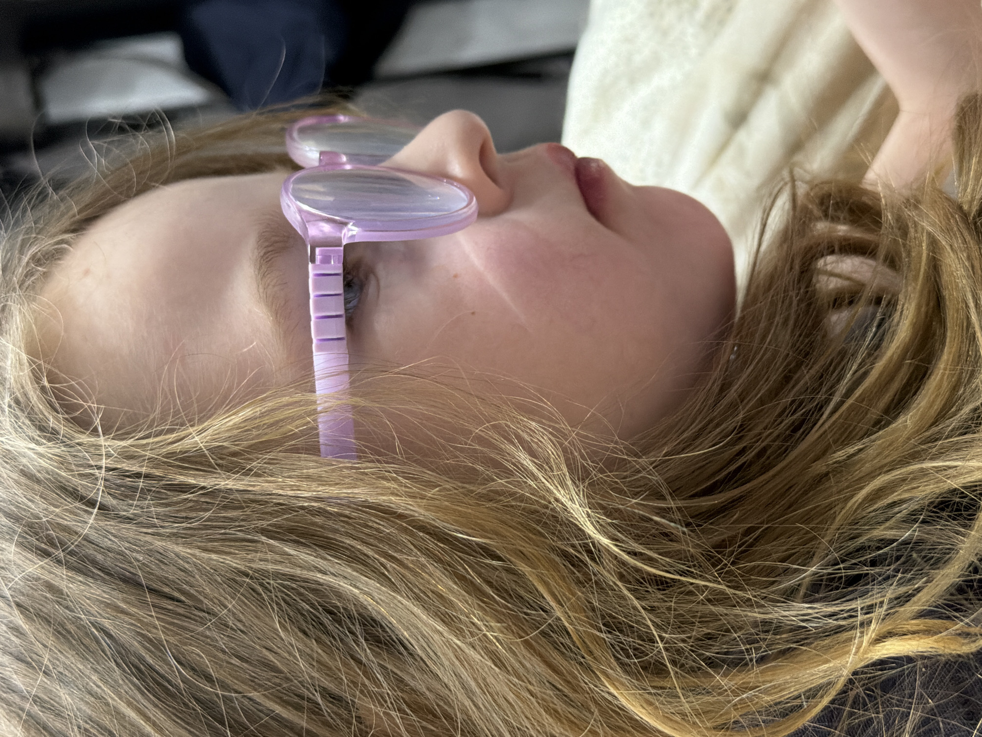 A close-up, ground-level shot features a young person lying down with their head turned towards the viewer. The person has fair skin, blonde hair, and is wearing lilac-colored glasses. The background consists of soft, white fabric or bedding and darker, blurred elements. There is no indication of the location being in the city of Chaikovsky, Russia, in this image.