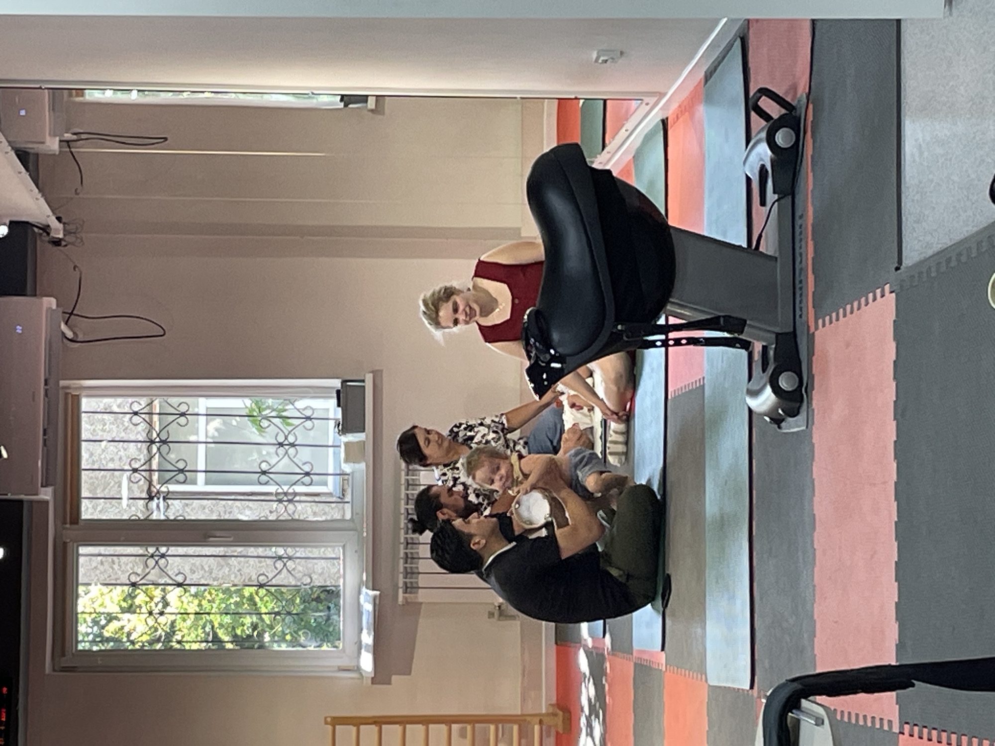 The image depicts an indoor environment, possibly a gym or a therapy center, located in Almaty, Kazakhstan. In the foreground, a black saddle-like exercise machine is prominently displayed on interlocking mats. A group of people are gathered on the mats, some seated and others holding a baby. The background features large windows with decorative grilles, revealing greenery outside, and wall-mounted air conditioning units. The scene suggests a class or a session involving exercise and childcare.