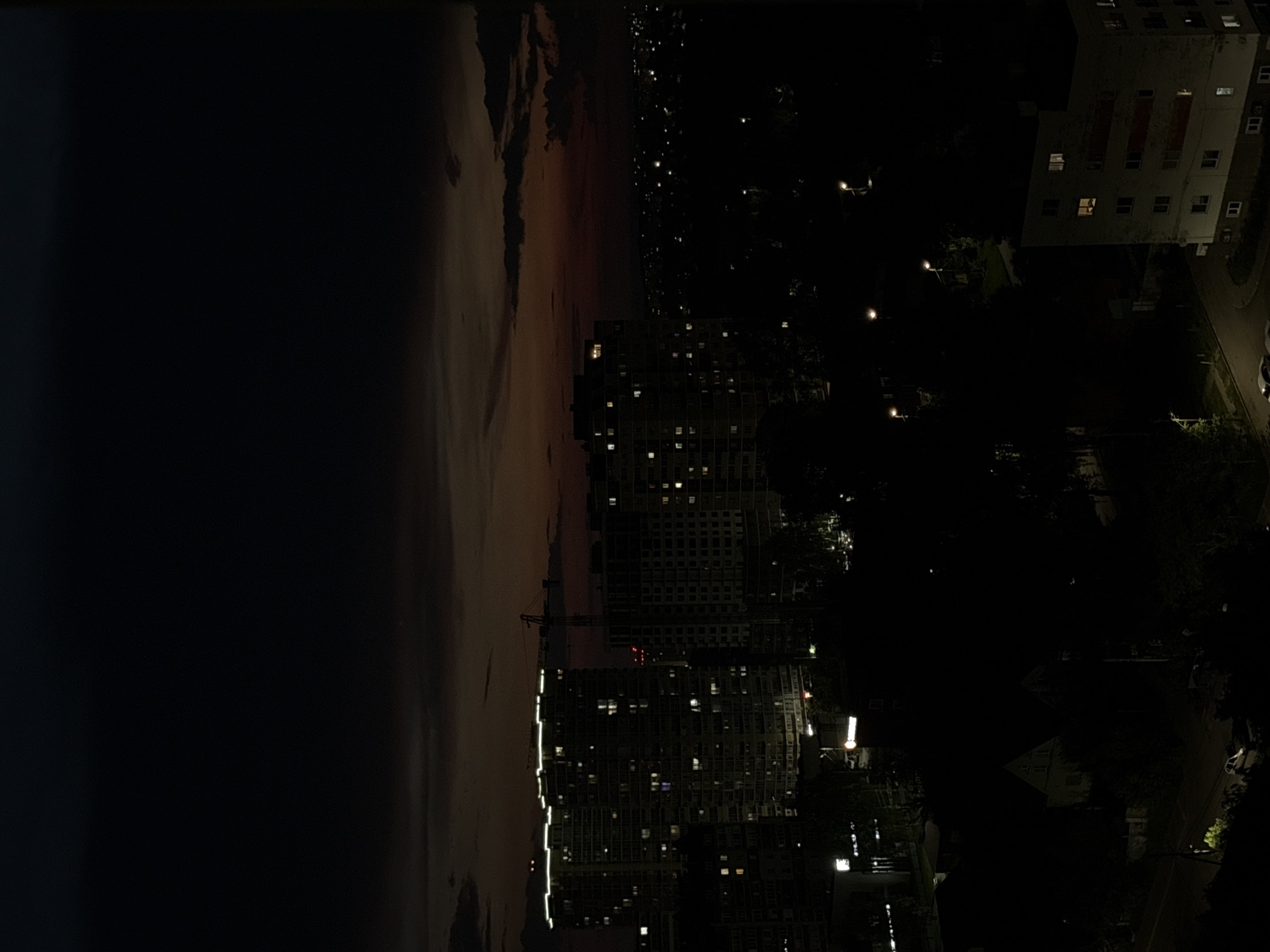 The image depicts a nighttime cityscape in Izhevsk, Russia. In the foreground, the silhouettes of trees and some smaller residential buildings are visible. In the midground, a cluster of tall, modern apartment buildings with illuminated windows dominates the scene. One building on the left features a bright, continuous band of light along its upper edge, possibly an architectural lighting feature. A construction crane is positioned behind these buildings, indicating ongoing development. The background shows a sprawling urban area with numerous scattered lights, suggesting more residential areas and perhaps some industrial or commercial zones. The sky is dark, with some diffused light from the horizon indicating the twilight or post-sunset period, and some wispy clouds are present. The overall impression is that of a typical urban environment at night, characterized by artificial light emanating from residential structures and streetlights, contrasting with the darkness of the sky and unlit areas. There are no discernible people or specific actions depicted in the image.