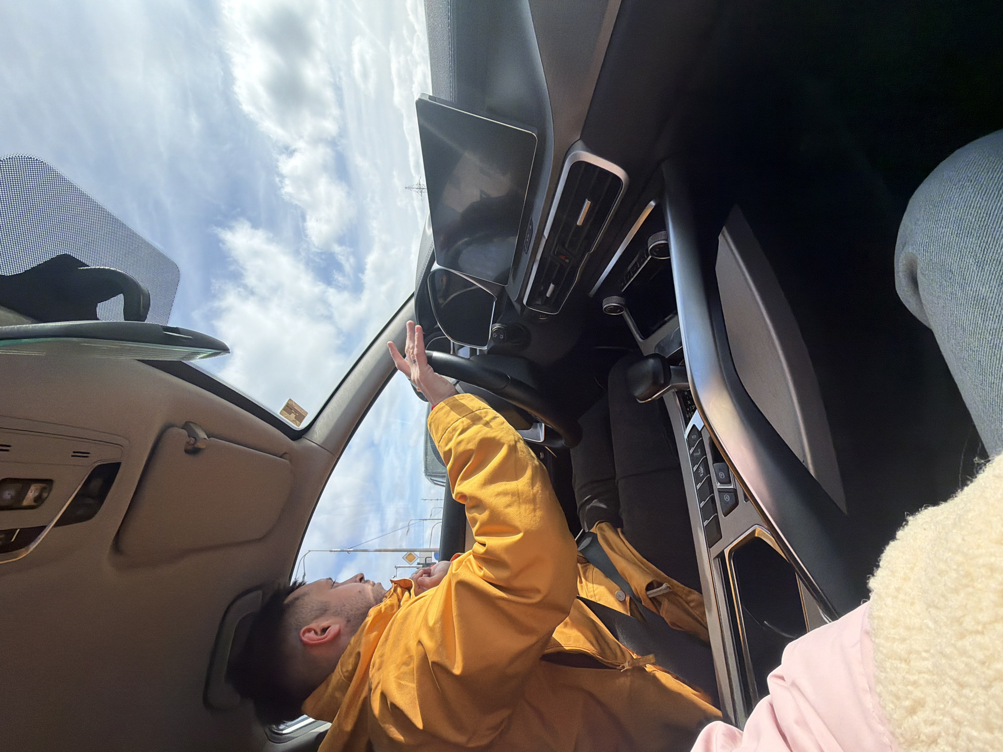 The image depicts the interior of a car. A person with short dark hair is visible in the driver's seat, wearing an orange jacket and facing towards the windshield. Their left hand is raised towards the steering wheel. The sky with clouds is visible through the windshield. The car's dashboard, center console, and other interior features are partially visible. The location of the image could not be determined.