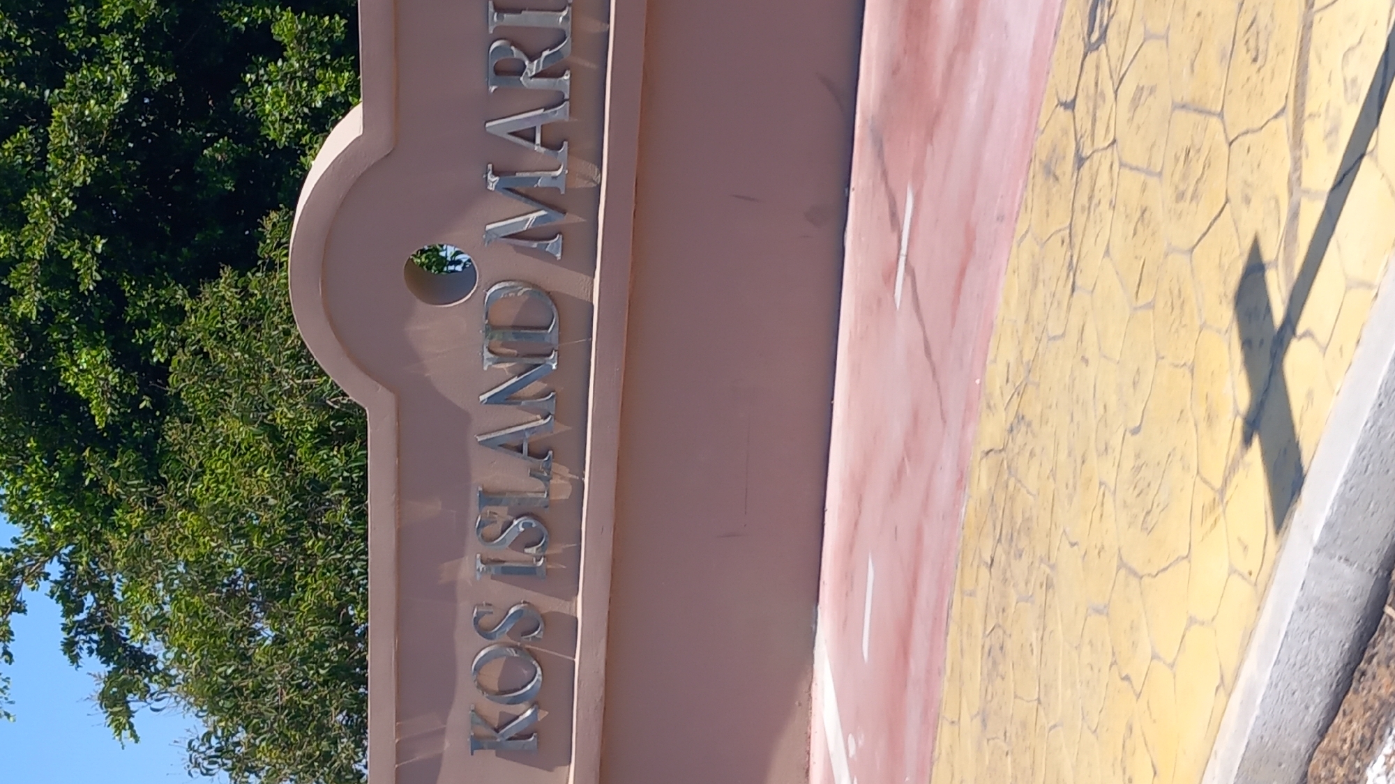 The image shows a sign that reads "KOS ISLAND MARINA". The sign is made of metallic letters attached to a pinkish-beige wall. Above the wall, green trees are visible against a bright blue sky. In the foreground, there is a path with a red section and a yellow section with a stamped pattern. A shadow of a cross-shaped object is cast on the yellow path. The scene suggests the entrance to a marina located in Kos, Greece, given the text on the sign.