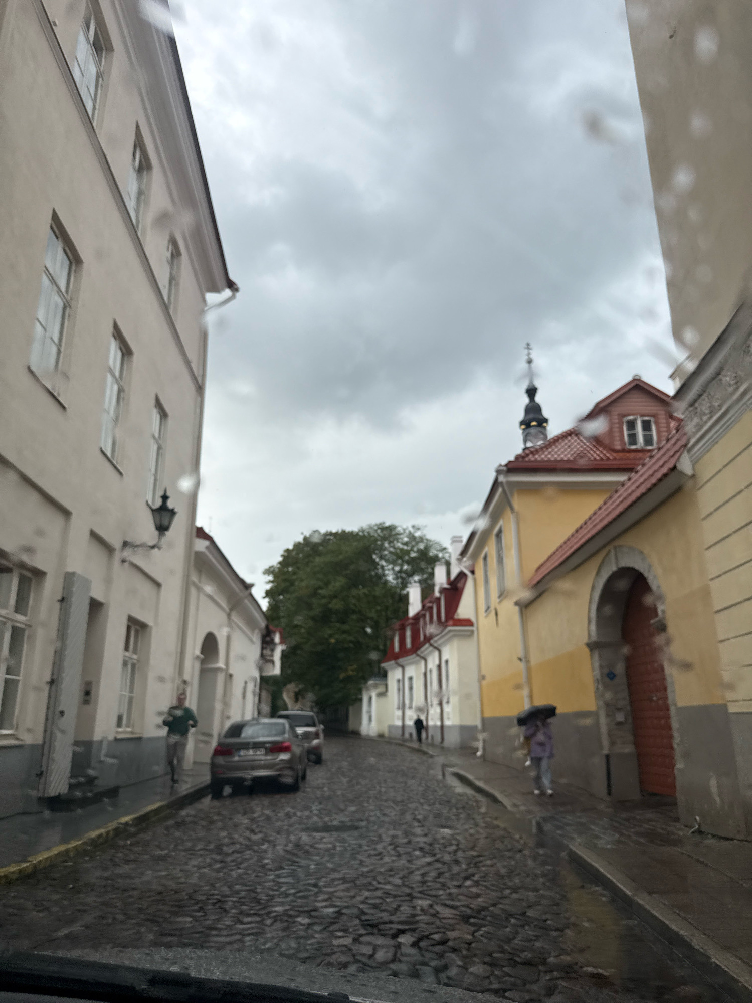 The image captures a view down a narrow, cobblestone street in Tallinn, Estonia, likely within the Old Town, characterized by its historical architecture. The sky is overcast and appears to be raining, as indicated by water droplets on the foreground lens and the wet appearance of the street.
On the left, a tall, light-colored building with multiple windows lines the street. A black, ornate street lamp is mounted on its facade. Further down on the left, a man in a green shirt and grey pants walks away from the camera. A silver sedan is parked on the left side of the street.
On the right, buildings feature facades in yellow and cream colors, with red-tiled roofs. One building has a distinctive arched doorway with a red door. Further down the street on the right, a person walks under a dark umbrella, wearing a light-colored jacket and grey pants.
A large, green tree is visible in the middle distance, and the silhouette of a church spire can be seen against the cloudy sky. The overall atmosphere is somber due to the weather conditions. The image is taken from a low vantage point, possibly from inside a vehicle.