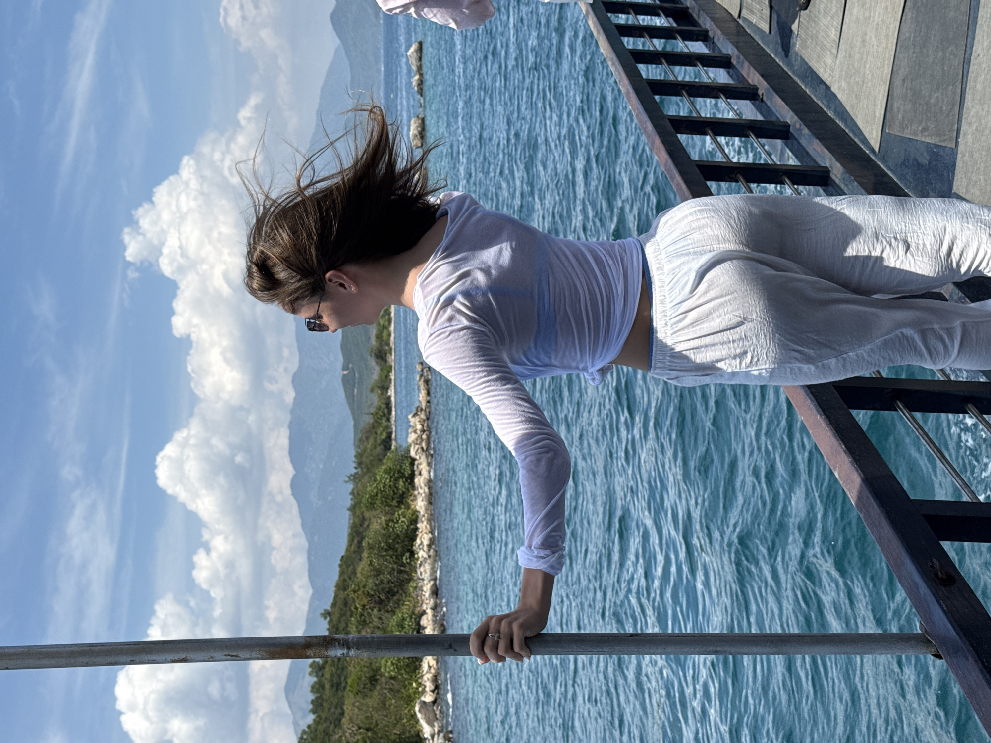 A young woman stands on a wooden pier in Nha Trang Ward, Vietnam. She is dressed in a white, long-sleeved top and white pants, her hair blowing in the wind. She wears sunglasses and leans forward, holding onto a metal railing. To the left, the turquoise sea stretches out to a shoreline with green vegetation and distant mountains under a blue sky with white clouds. A wooden ladder is visible to the right, suggesting the pier's edge. The scene suggests a sunny day by the water.