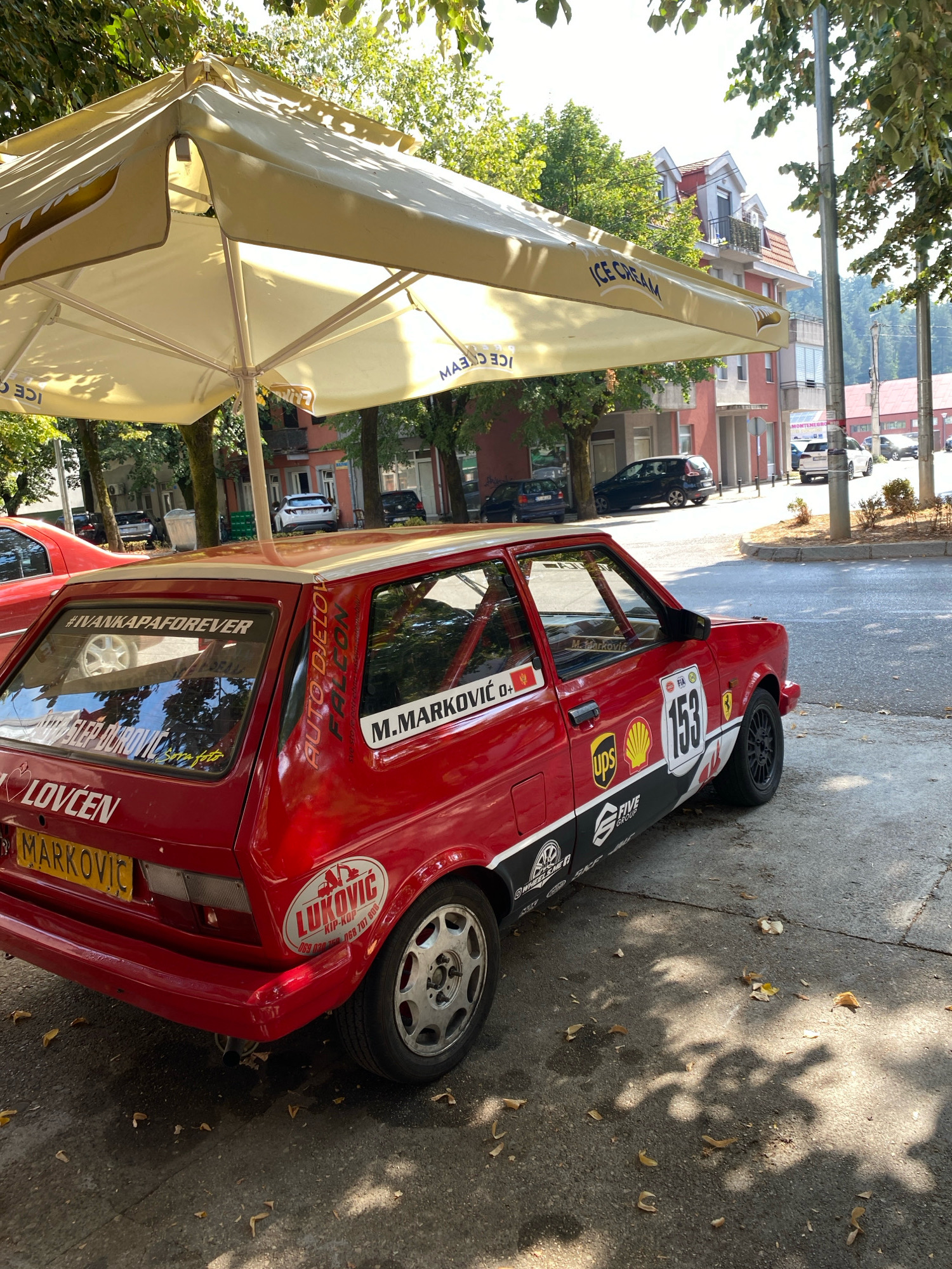 An exterior, medium shot captures a red Yugo car with racing decals parked on a paved street in Cetinje, Montenegro. The car is positioned at an angle, with its rear facing the viewer. The car has various stickers, including "LOVCEN MARKOVIC", "#IVANKAPAFOREVER", and "M. MARKOVIC". The car is partially shaded by a large, cream-colored umbrella advertising "ICE CREAM". In the background, there are buildings with red roofs, trees, and other cars on the street. The sky is bright and appears to be a clear day. The ground is asphalt, with scattered fallen leaves. The overall scene depicts a typical urban environment on a sunny day.