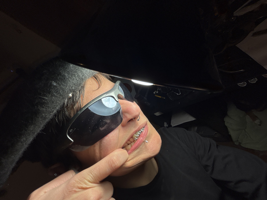 The image depicts an individual in a dark interior environment, likely a vehicle. The subject wears dark, wraparound sunglasses with silver-grey frames, a dark long-sleeved top, and a dark beanie. Metallic silver grillz cover the subject's upper teeth. A silver septum ring is visible below the nose, a silver vertical labret piercing on the lower lip, and two additional silver piercings are present on the upper left ear. The subject's left index finger pulls down the lower lip, exposing the lower teeth and the labret piercing. A prominent vertical light source illuminates the right side of the scene, reflecting brightly in the right sunglass lens. The background is dark and out of focus, showing indistinct forms consistent with a vehicle's dashboard and a light-colored soft object, possibly upholstery, at the bottom of the frame. No identifying features are present to determine a specific location within Sofia, Bulgaria.
