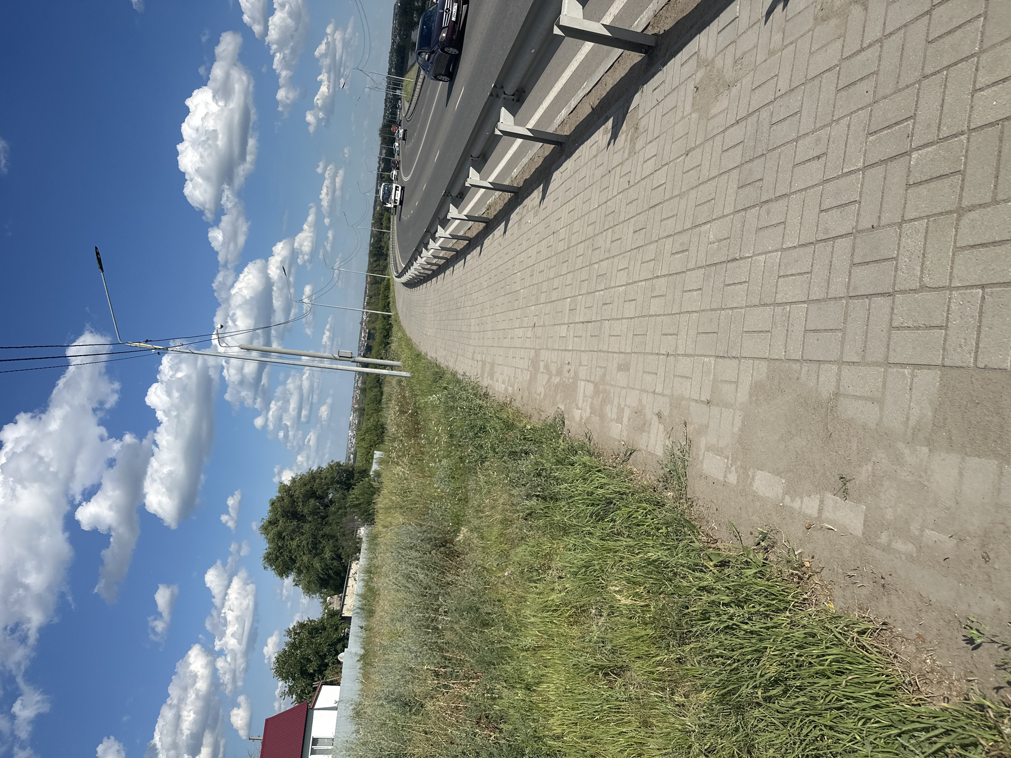 This image shows a paved sidewalk running alongside a highway. The sidewalk is made of light grey rectangular pavers laid in a basketweave pattern. To the left of the sidewalk is an area of tall, green grass and weeds, with a white building with a red roof and a grey fence in the background. The highway is to the right, with several cars and a truck driving on it. A metal guardrail separates the sidewalk from the highway. There are several streetlights along the highway. The sky is blue with scattered white clouds. The scene appears to be an urban or suburban environment, possibly on the outskirts of a city. The location is identified as Kostanay, Kazakhstan.
