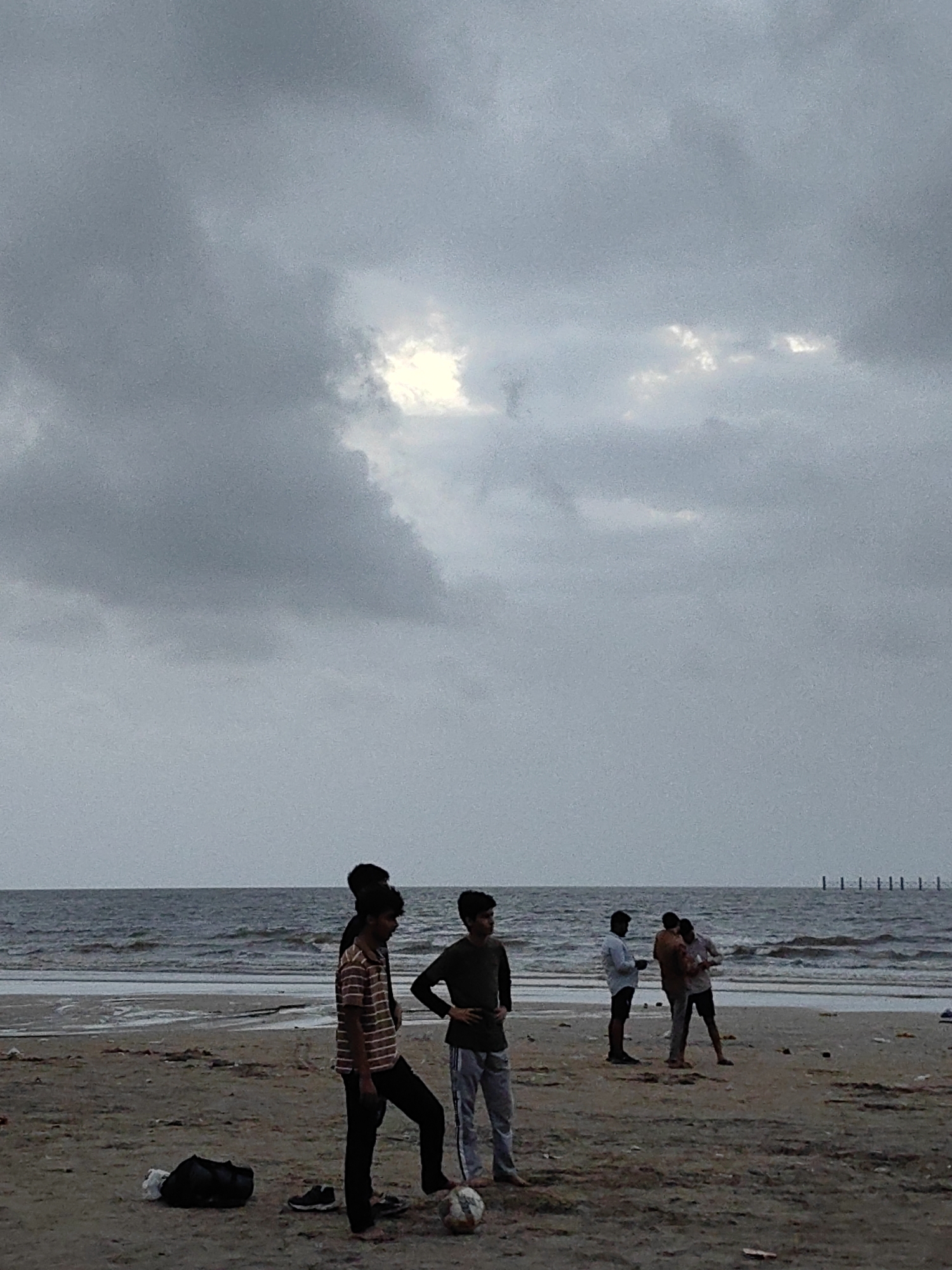 The image captures a group of young men on a beach in Mumbai, India, under a cloudy sky. The scene depicts a sandy beach with the ocean in the background, characterized by choppy waves and a distant horizon marked by a line of pilings. Several individuals are visible: one man in the foreground on the left stands with a striped t-shirt and dark pants, with a soccer ball at his feet. To his right, another man in a dark shirt and light pants stands facing away. Further back, a group of three men are engaged in conversation or looking at their phones. The atmosphere appears overcast, suggesting a pre-monsoon or post-monsoon day. The beach itself shows signs of debris scattered on the sand. The overall impression is one of casual recreation on a coastal area.