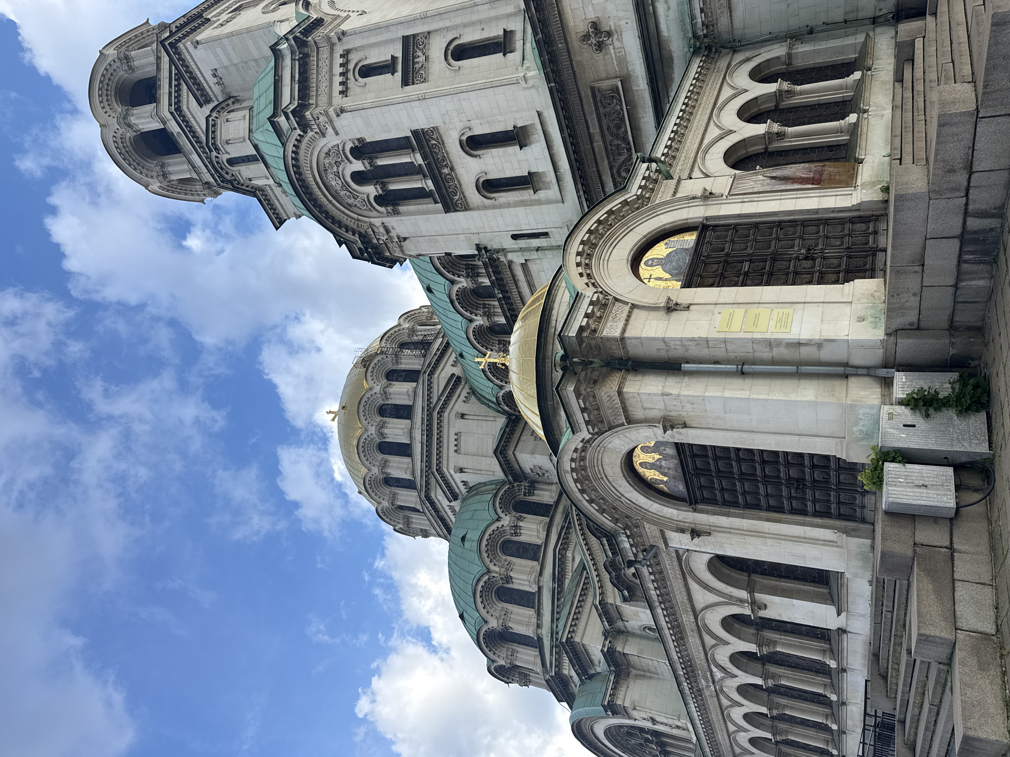 The image shows an upward-angled view of the Alexander Nevsky Cathedral in Sofia, Bulgaria. The building is constructed of light-colored stone and features domes with gold and green roofing. Arched windows and ornate carvings adorn the facade. Above the building is a blue sky with scattered white clouds. The image appears to be taken from street level.
