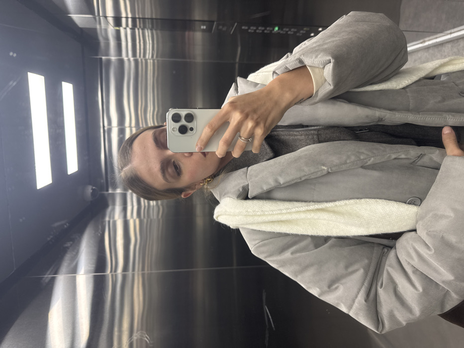 An adult female is captured in a mirror selfie within an elevator in the city of Unknown, Unknown. She stands facing forward, holding a light-colored smartphone with a multi-lens camera in her right hand, positioned to photograph her reflection. Her gaze is directed towards the phone's screen. She wears a light grey puffer-style coat over a darker grey garment and a cream-colored top. A cream-colored knitted scarf is draped around her neck and shoulders. A silver-toned ring is visible on her right ring finger, and she wears gold-colored hoop earrings. The elevator interior features brushed metallic walls with horizontal striations. Two rectangular overhead lights are illuminated, and a small spherical security camera is visible mounted on the ceiling in the reflection. A portion of an elevator control panel is visible on the right wall.