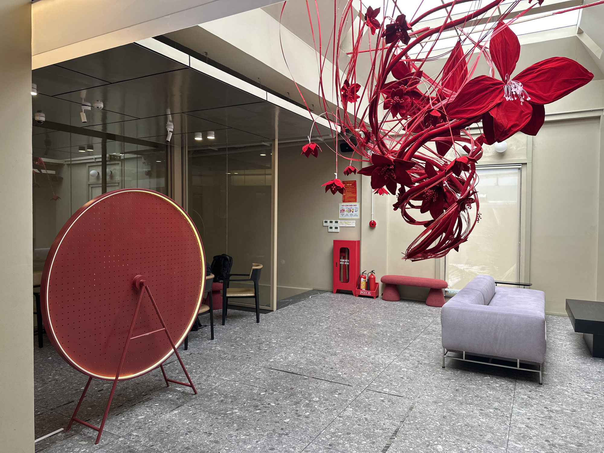The image depicts an indoor space, possibly a lobby or exhibition area, featuring a large red circular installation with a light ring and a stand. Suspended from the ceiling is an elaborate red floral art installation with multiple large, velvet-like flowers and delicate tendrils. To the right, a light purple sofa with silver legs sits on a speckled grey floor. Adjacent to the sofa is a dark rectangular coffee table. Further to the right and behind the sofa is a muted red bench with rounded legs. In the mid-ground, a bright red fire extinguisher cabinet is visible, with two smaller red fire extinguishers inside. Near the fire cabinet, a sign is affixed to the wall. Chairs with black frames and wooden seats are placed in the background. The overall impression is one of modern interior design with a focus on vibrant red accents. The location of this scene is in Đà Nẵng, Vietnam.