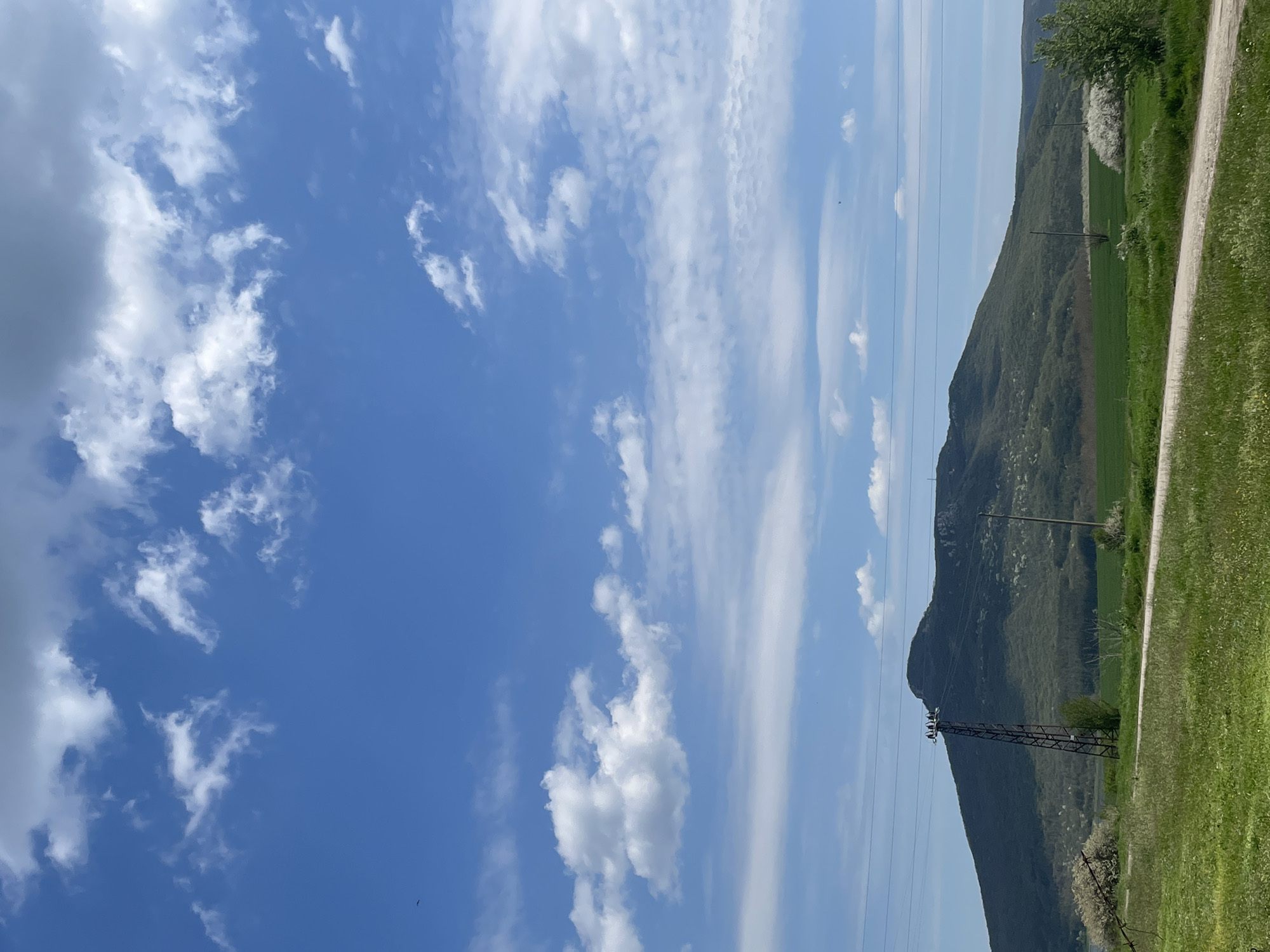 The image shows a landscape on a sunny day in Buhovtsi, Bulgaria. In the foreground, there is green grass on the right side of the image, as well as a dirt road. A power line is visible, supported by an electric pylon. Behind the pylon, there is a green mountain covered in vegetation. The sky is blue with white clouds.